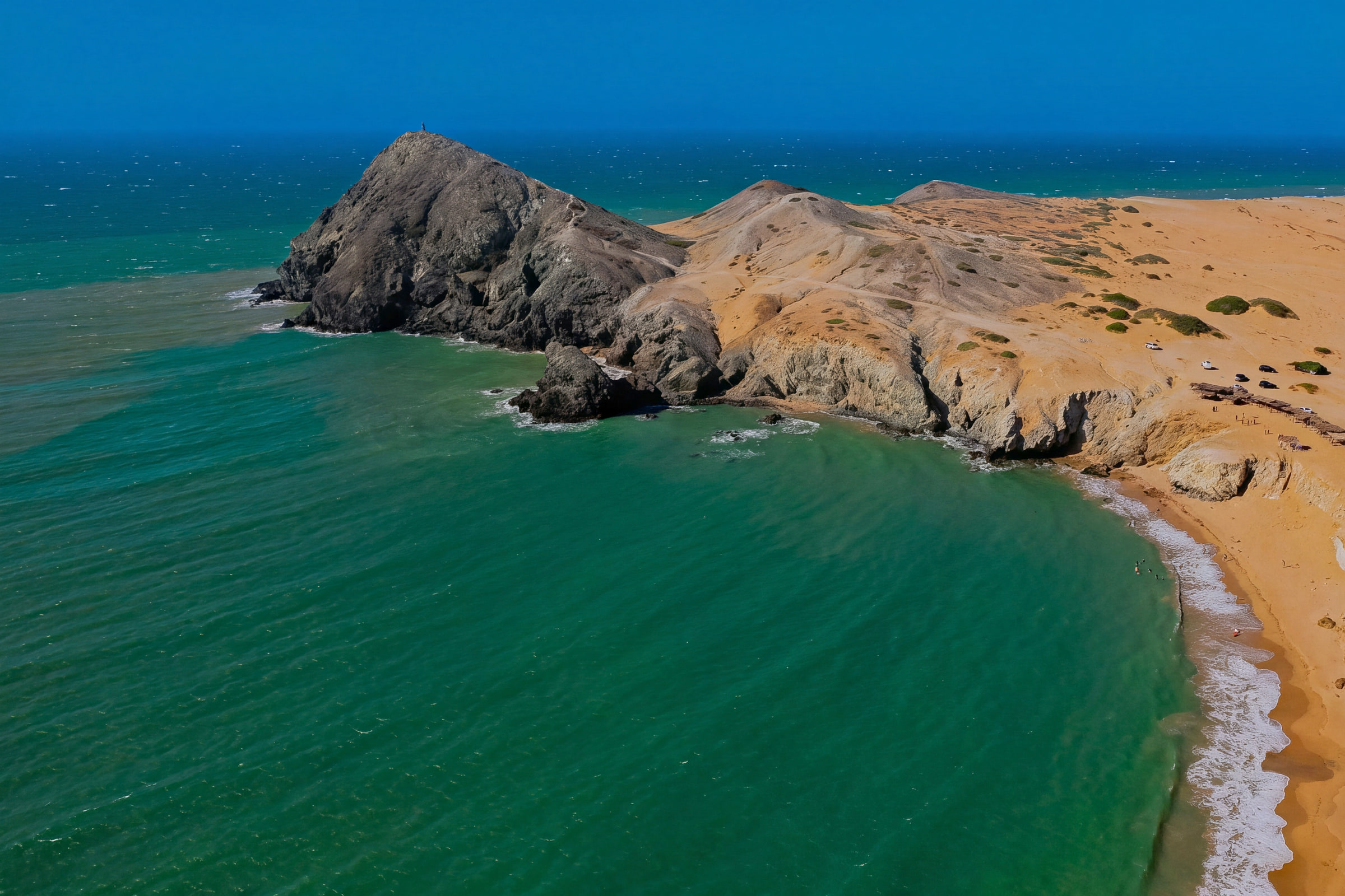 Aerial view of the Pilón de Azúcar, a rocky hill rising from green water, with Playa del Pilón on the right, in Cabo de la Vela, La Guajira.