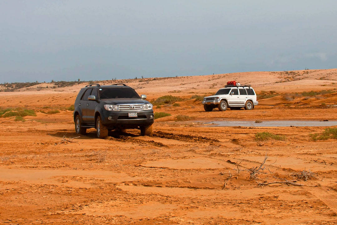 Dos camionetas 4×4 cruzando una planicie de tierra roja con charcos en el desierto de La Guajira, bajo un cielo nublado.