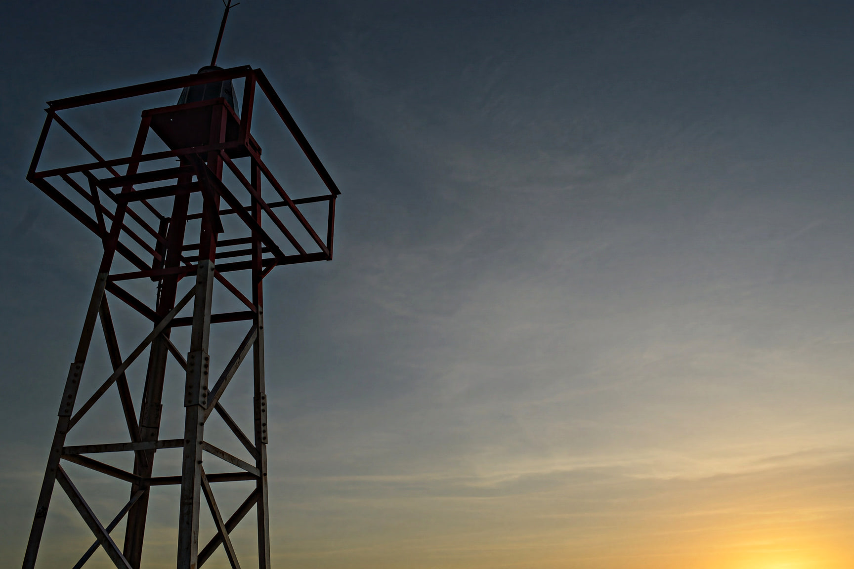 Silueta de la torre del faro del Cabo de la Vela al atardecer, con el sol a punto de ponerse sobre el mar.