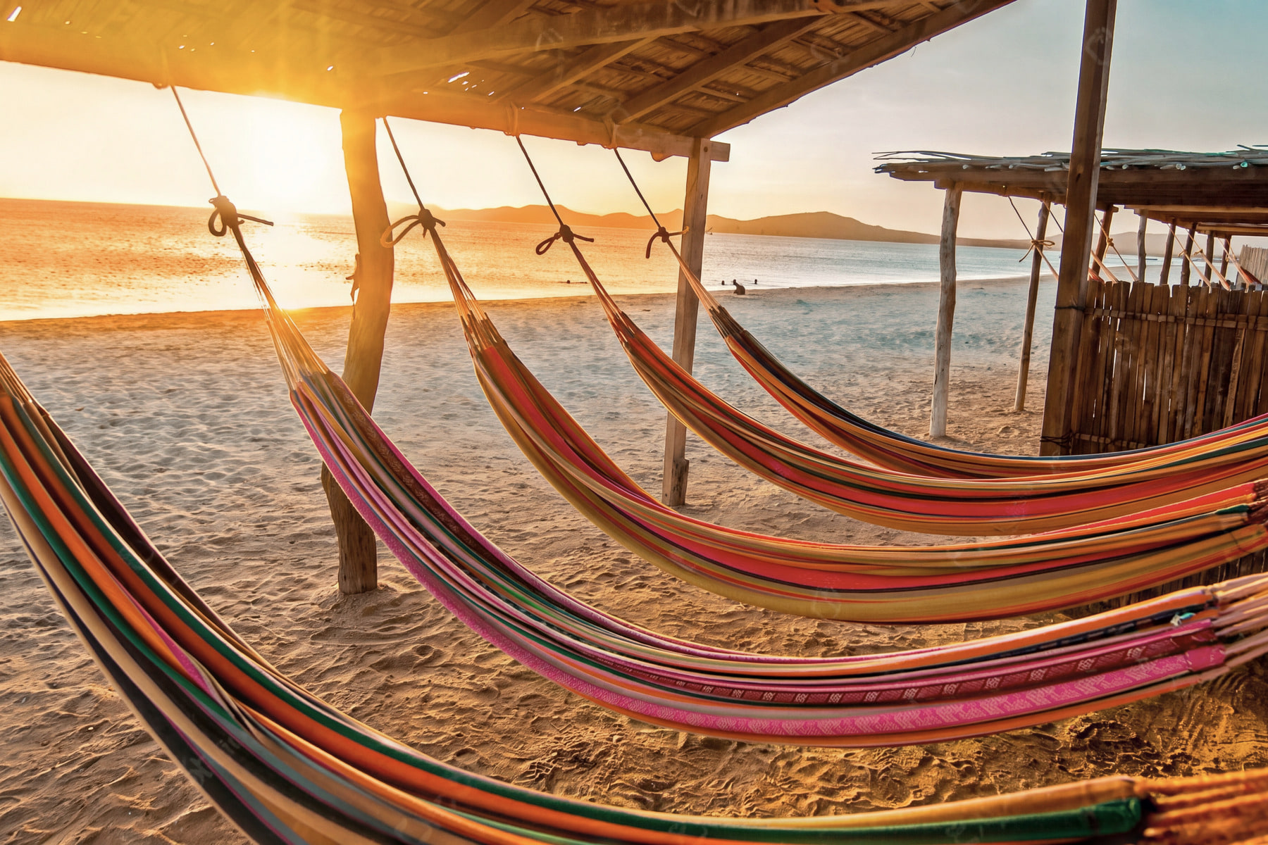 Colorful chinchorro hammocks under a palm shelter facing the sea at sunset, with the low sun between the posts, in Cabo de la Vela.