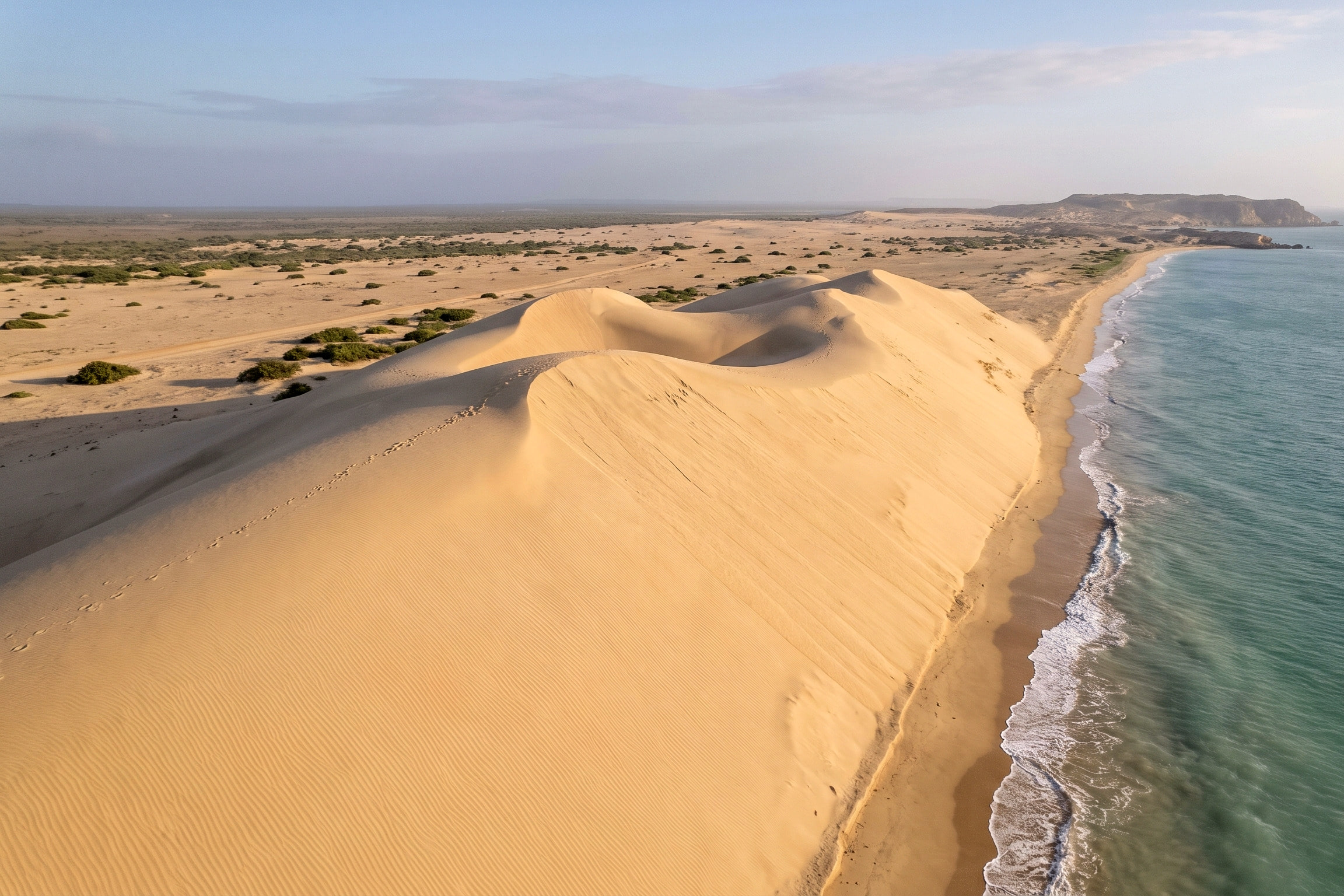 Vista aérea de las dunas de Taroa: una duna alta de arena clara baja hasta una playa estrecha con oleaje, junto al mar Caribe, en la Alta Guajira.