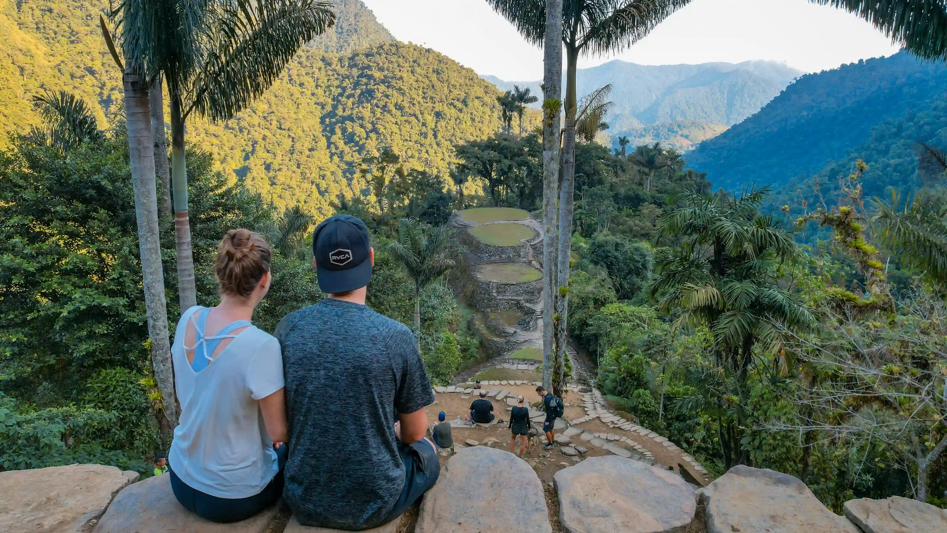 A couple sitting with their backs to the camera on a stone wall facing the terraces of Ciudad Perdida (Teyuna), with tall palms and mountains.