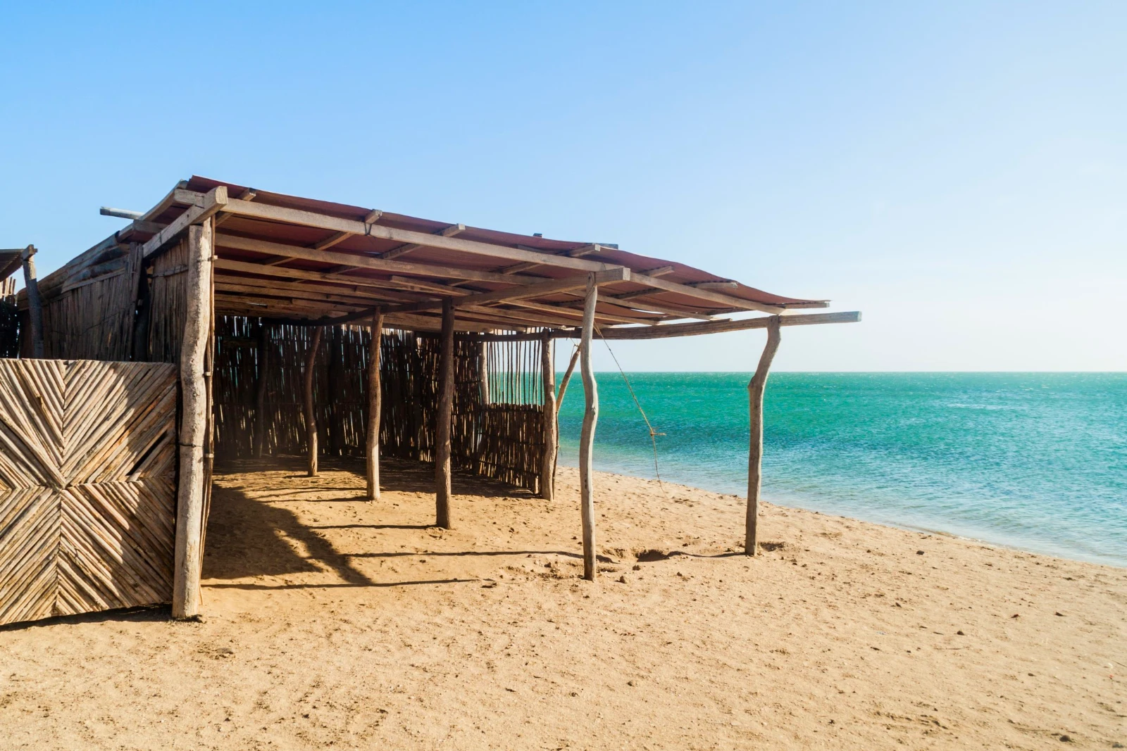 Enramada en perches et lattes de bois sur une plage de sable, mer turquoise à droite et ciel dégagé, à Cabo de la Vela.