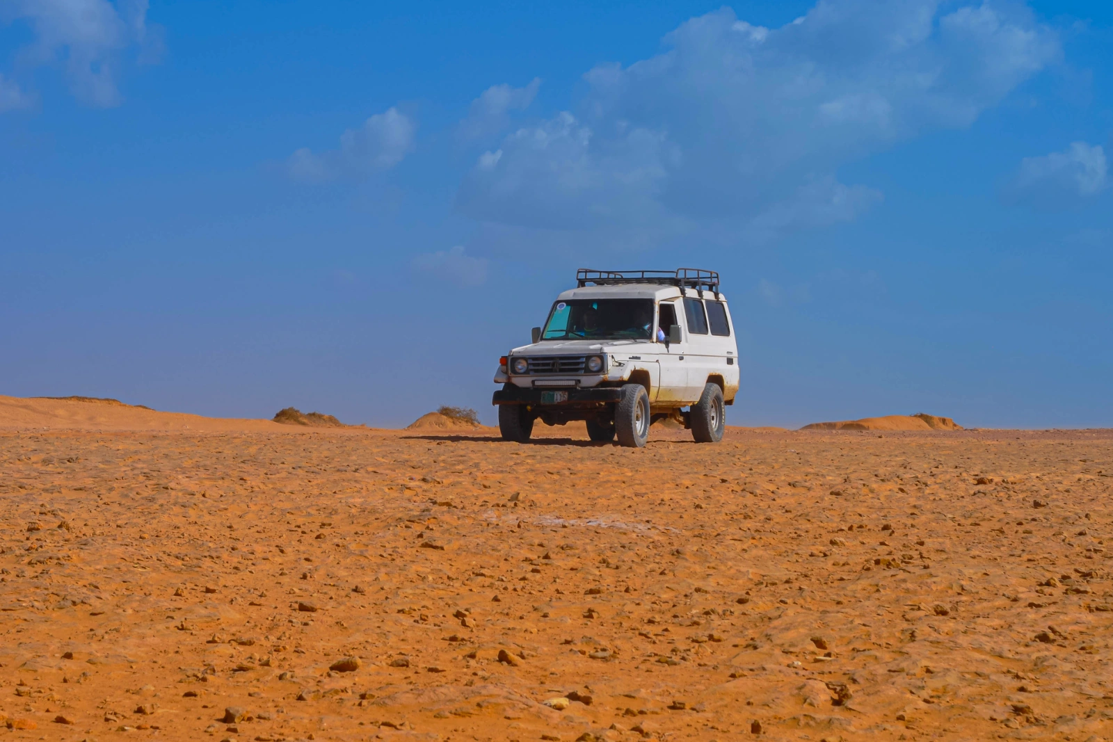 A white 4×4 with a roof rack stopped on an ocher desert plain of earth and stones, under a deep blue sky.