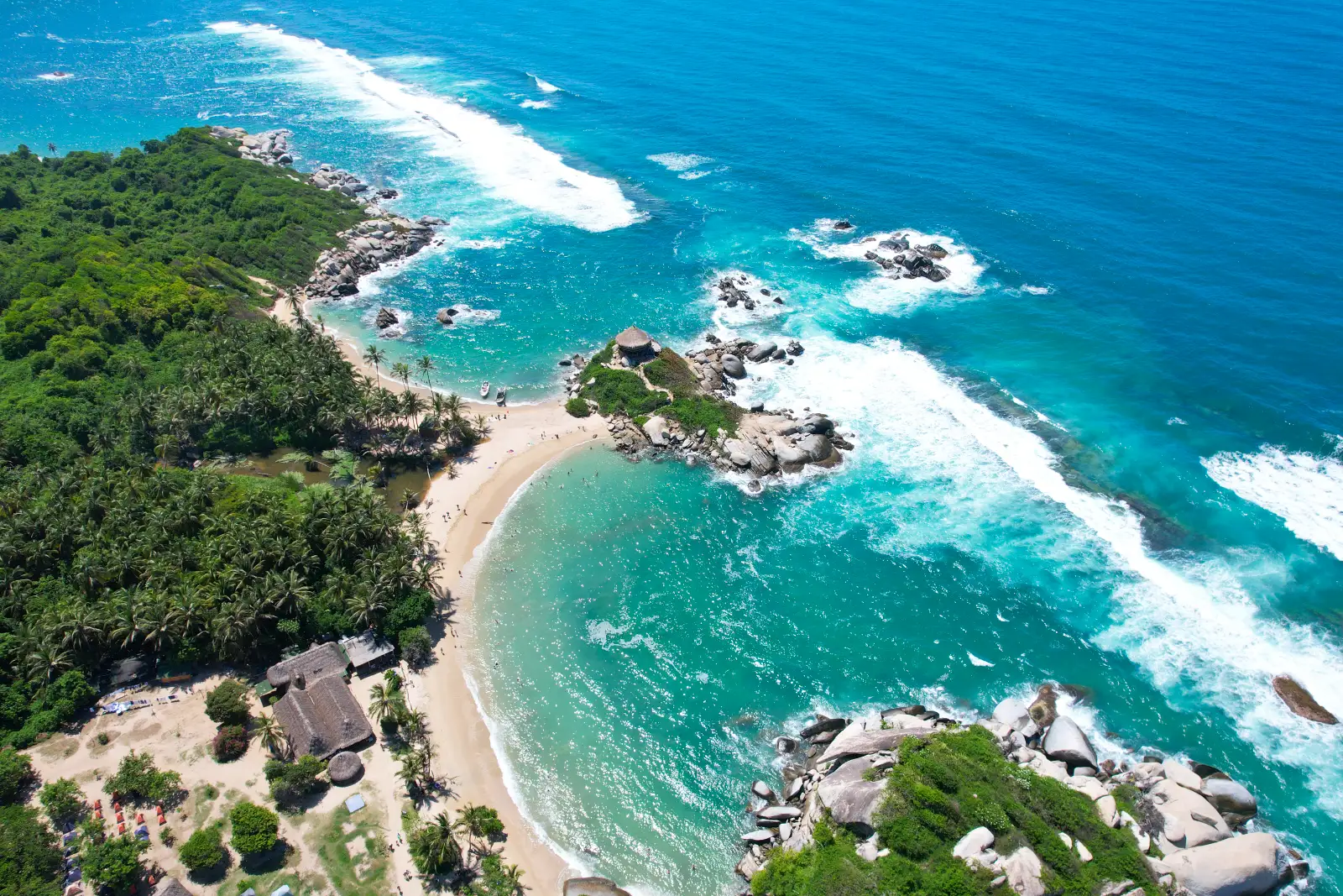 Vista aérea del Cabo San Juan del Guía: doble playa con palmas y cabañas, el islote con el kiosco y el oleaje blanco del mar abierto.