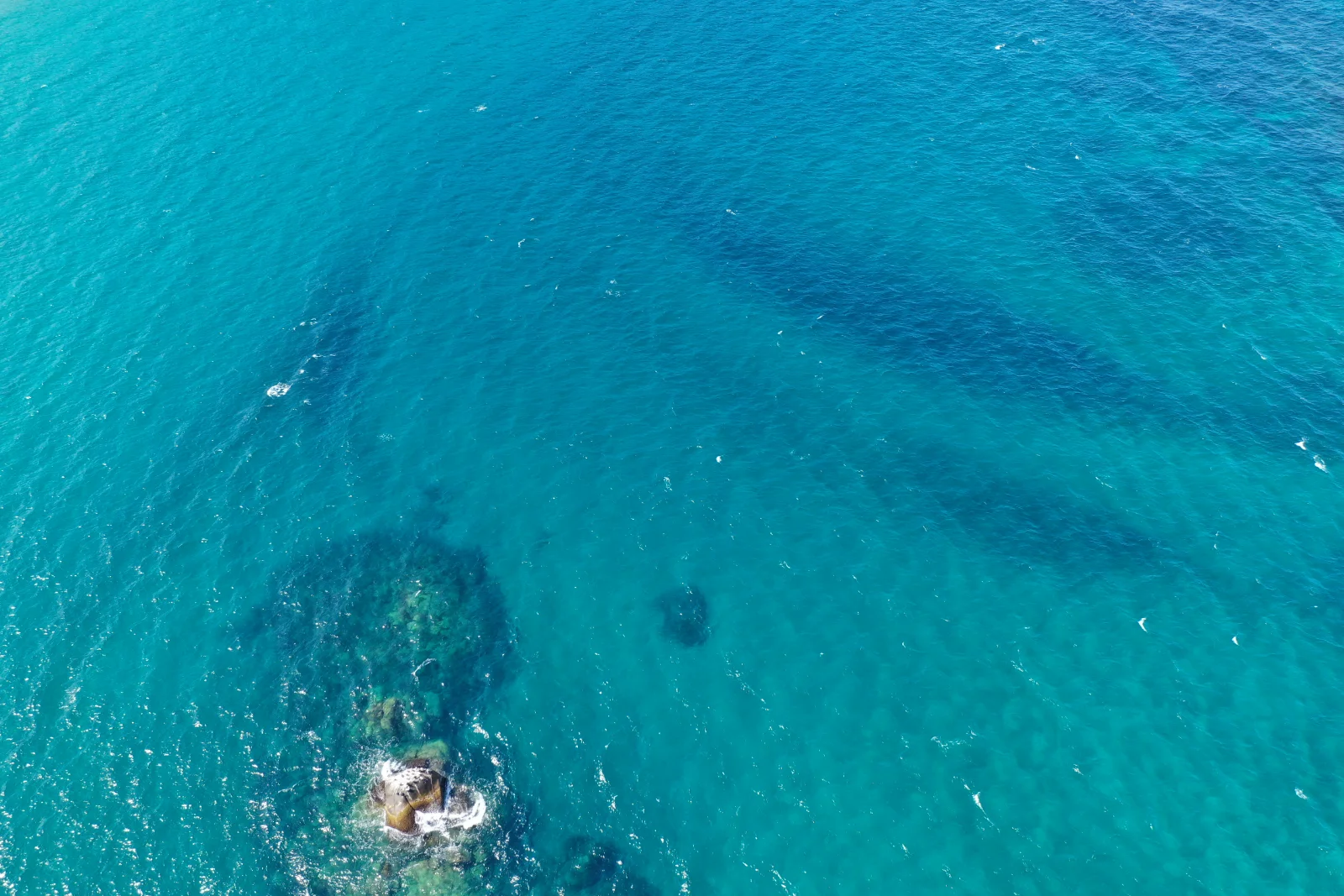 Vista cenital de agua turquesa con una roca que asoma y un parche de arrecife sumergido, frente a la costa del Parque Tayrona.