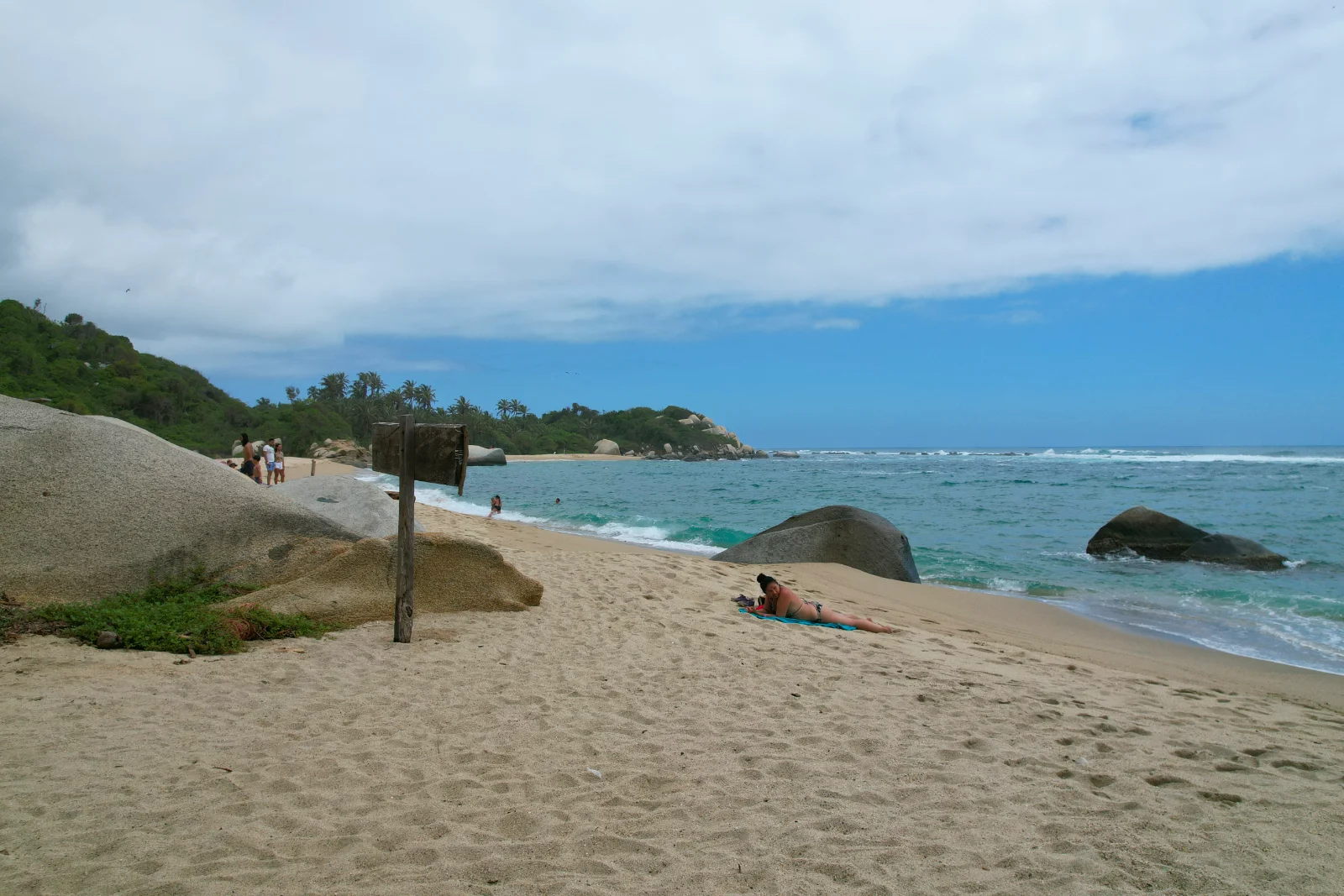 Playa de arena con bloques de piedra redondeados, un poste de letrero de madera y una persona tendida a lo lejos, bajo un cielo nublado.