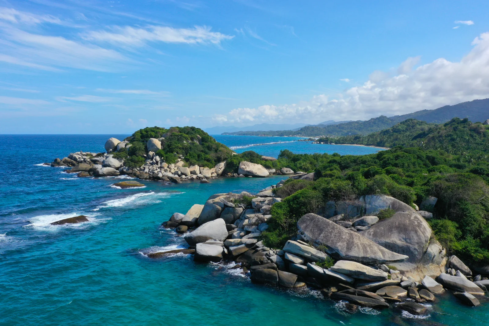 Vista aérea de islotes de grandes bloques de piedra con bosque, rodeados de mar azul, con la costa del Parque Tayrona y montañas detrás.