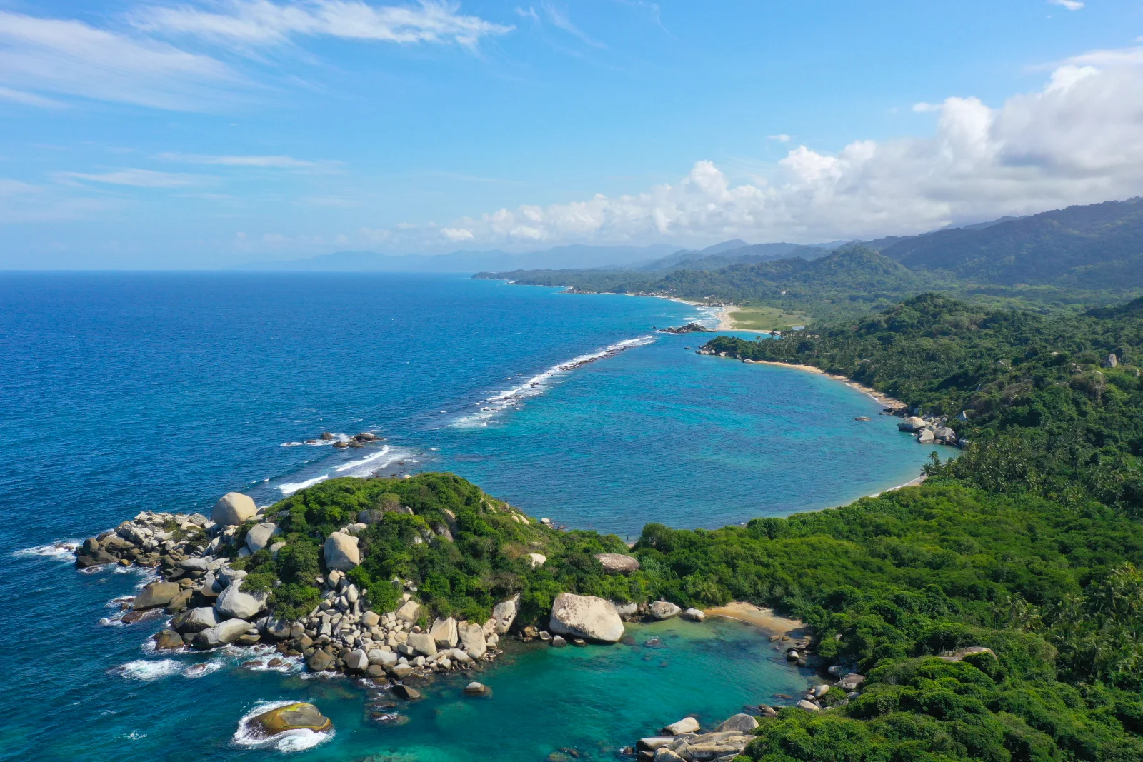Vista aérea de una punta rocosa con bloques de piedra, bahías de agua turquesa y playas largas hacia las montañas del Parque Tayrona.