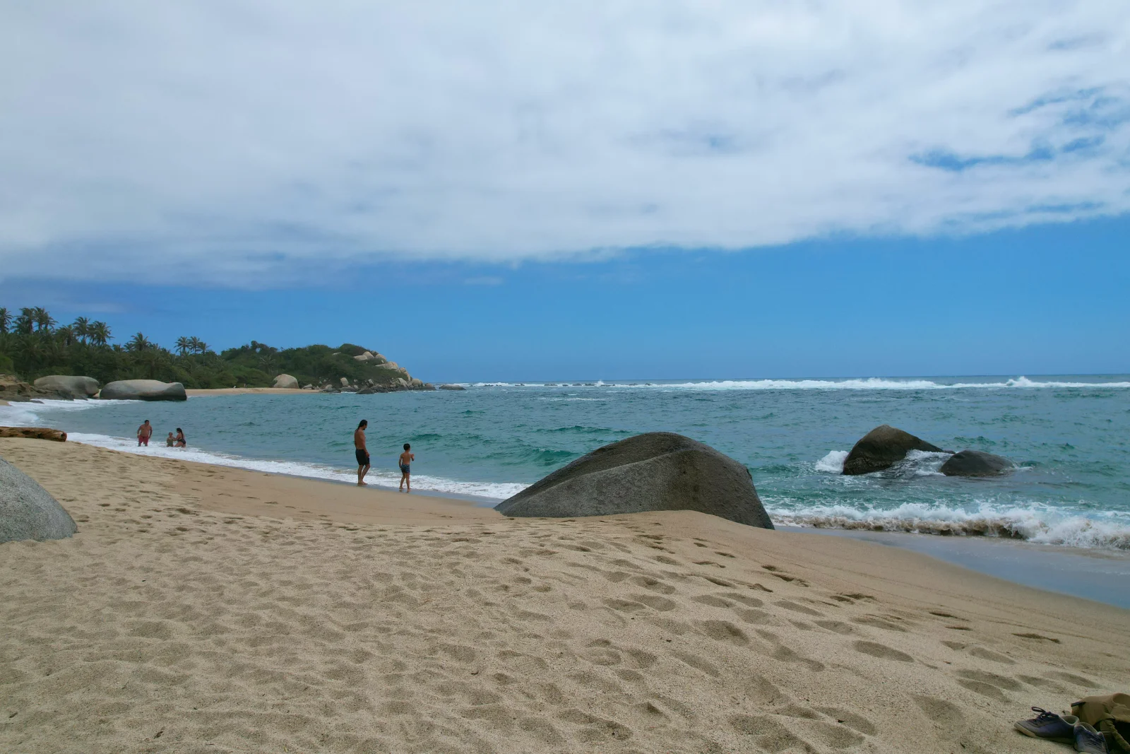 Playa de arena con huellas y un bloque de piedra redondeado en el agua, con algunas personas lejanas en la orilla y un cielo nublado.