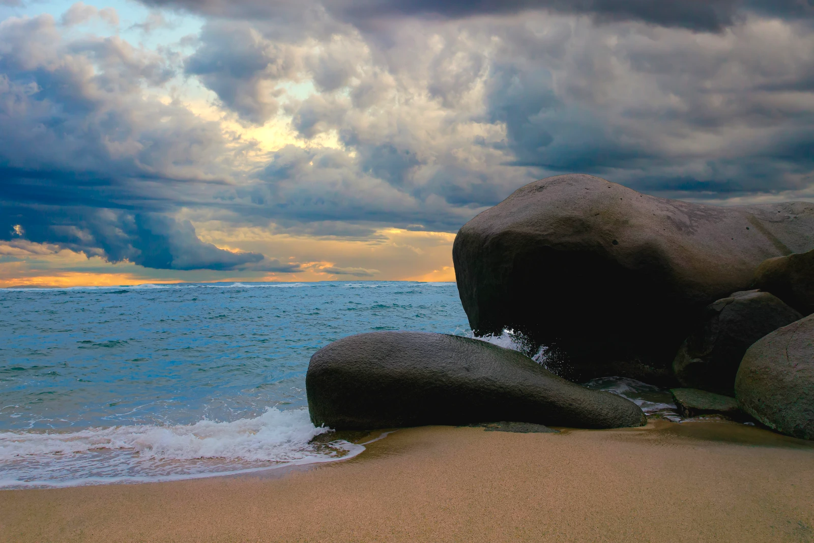 Grandes rocas oscuras y redondeadas en una playa al anochecer, con olas suaves, nubes densas y una franja de luz naranja en el horizonte.
