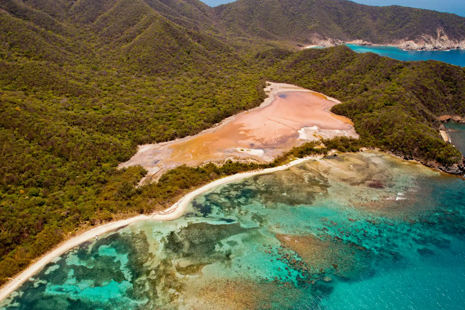 Vue aérienne d'une baie de Gairaca avec une lagune salée rose derrière la plage, un récif turquoise dans l'eau et des collines boisées.