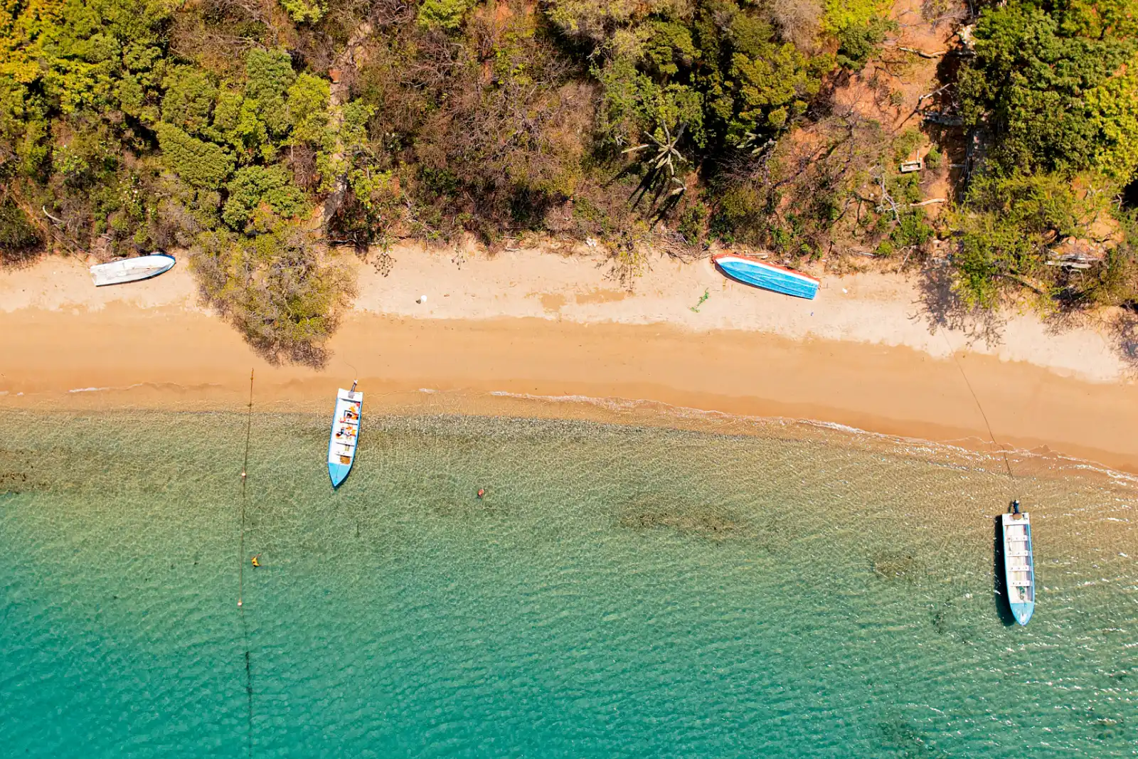 Vue zénithale d'une plage de sable avec trois petites barques, une eau turquoise peu profonde et la lisière de la forêt sèche, à Gairaca.