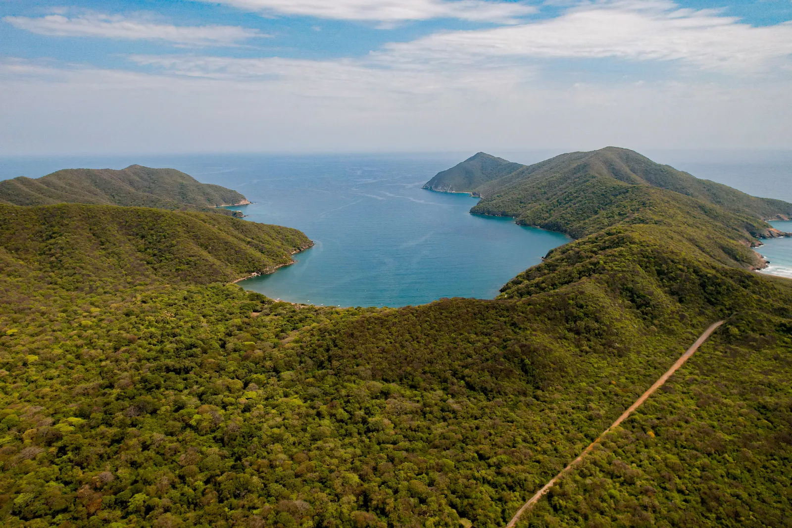 Vue aérienne de collines boisées autour d'une baie d'eau bleue, avec des caps au fond et un chemin de terre à droite.