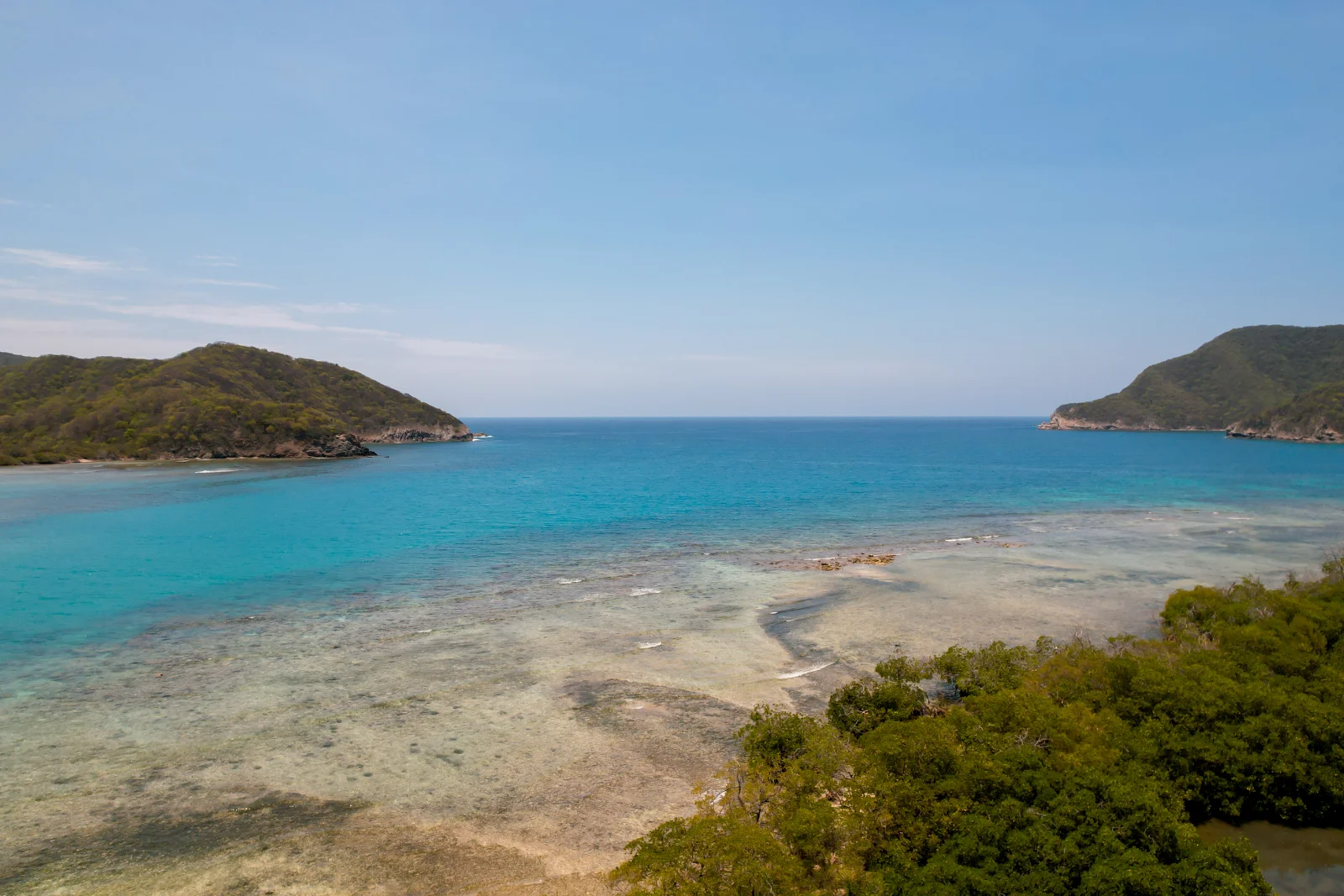 Baie peu profonde au récif visible, mangrove verte au premier plan, entre deux caps boisés sous un ciel bleu pâle.