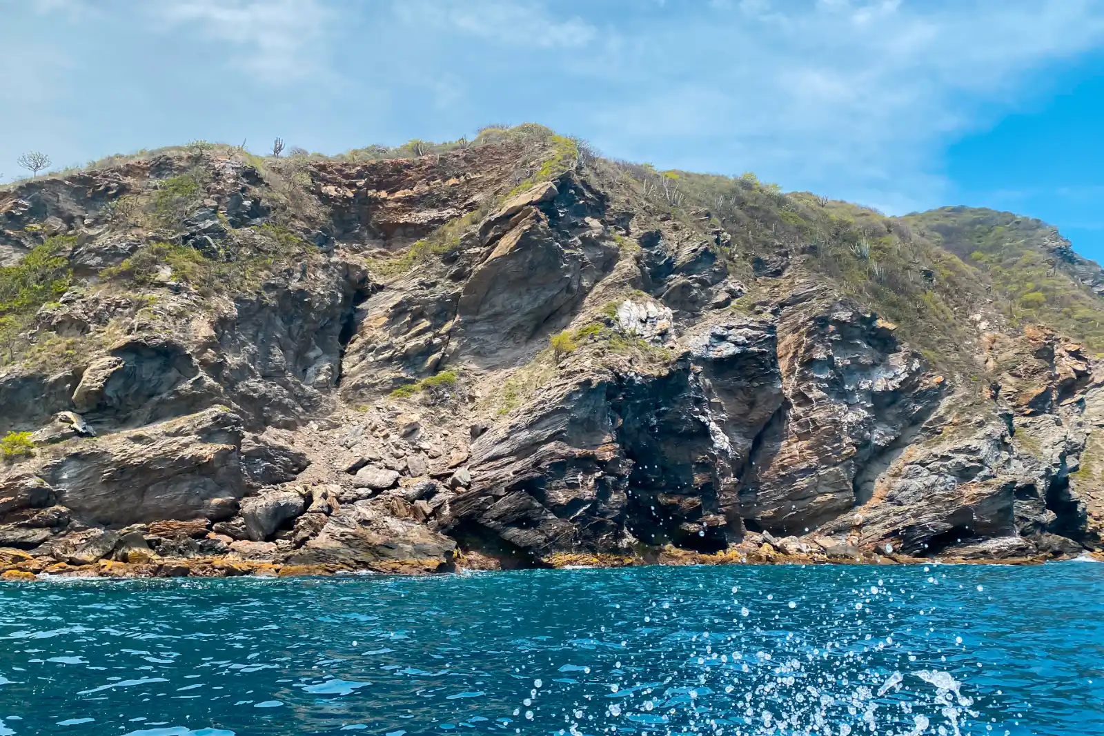 Falaise rocheuse veinée couverte de broussailles sèches vue de la mer, avec une eau bleue agitée au premier plan et un ciel nuageux.