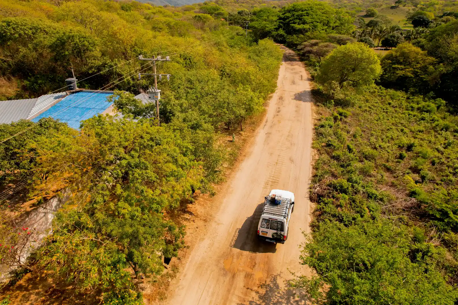 Vue aérienne d'un 4×4 blanc sur un chemin de terre à travers la forêt sèche, avec un bâtiment au toit bleu à gauche.