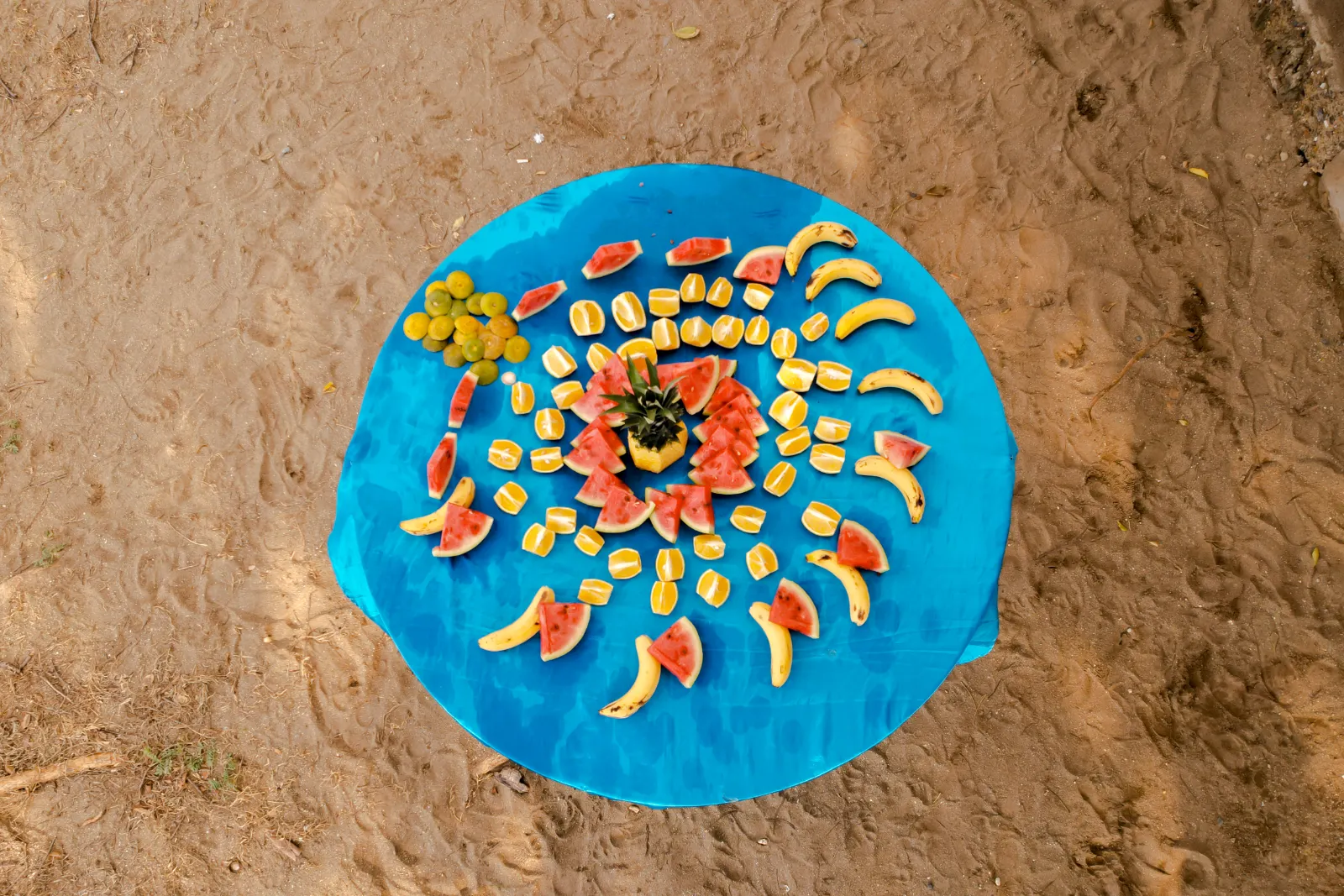 Vue zénithale de fruits coupés disposés en spirale, pastèque, banane et ananas, sur une nappe bleue ronde posée sur le sable.