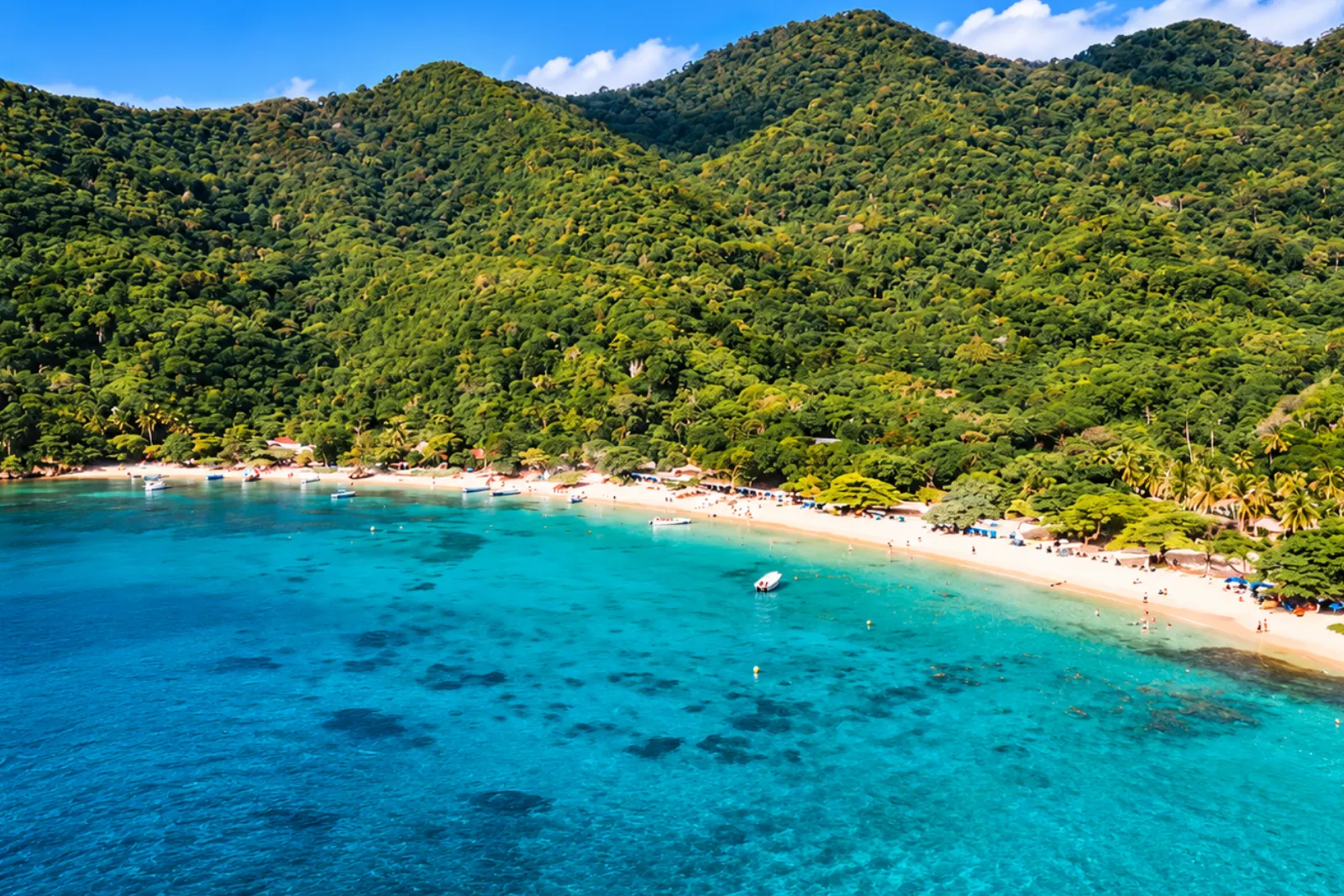 Vue aérienne de Playa Cristal : eau turquoise parsemée de récifs, bateaux sur le rivage et collines vertes boisées derrière la plage.