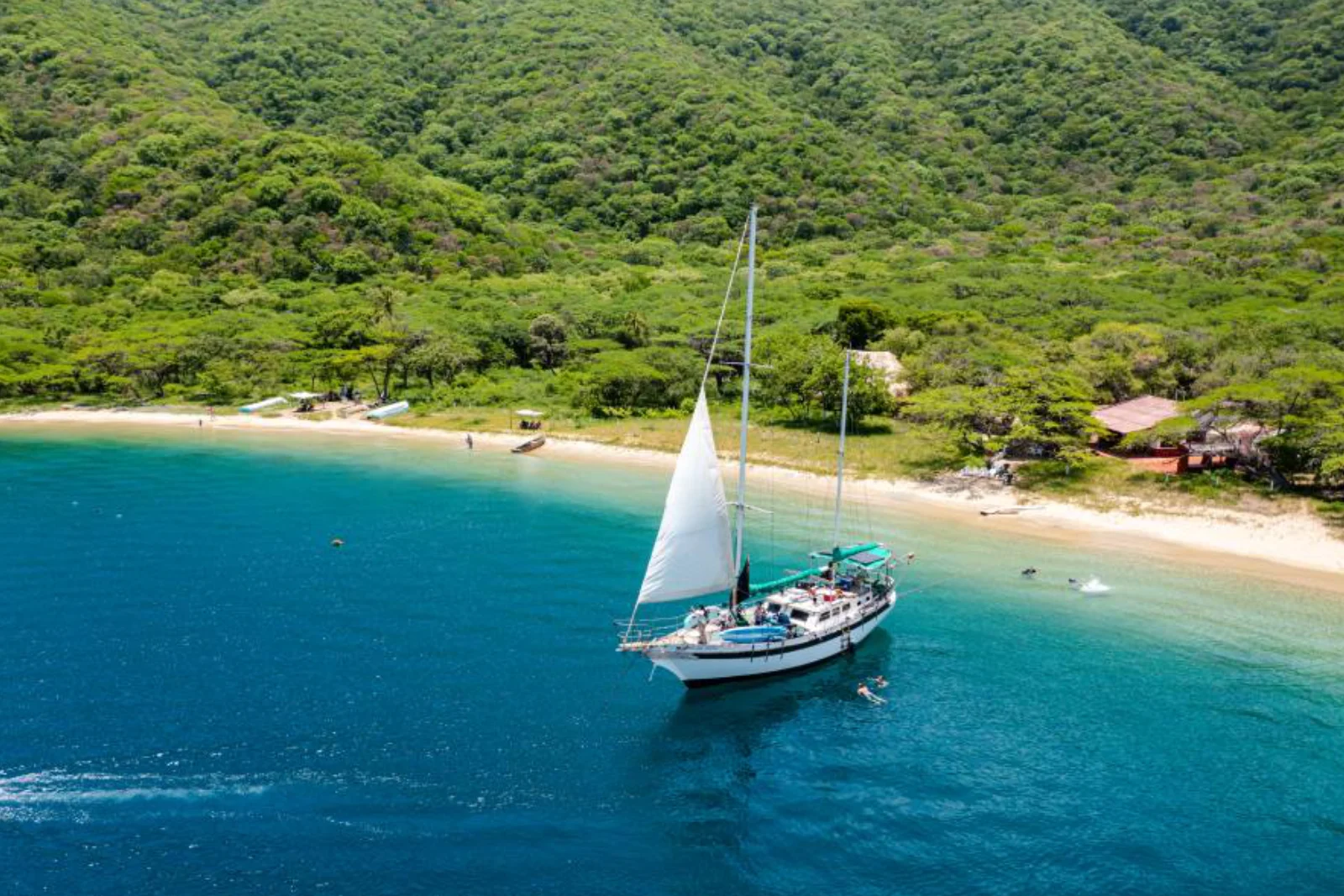 Voilier blanc, voile hissée, au mouillage devant une plage adossée à des collines vertes, quelques personnes nageant près de la coque.