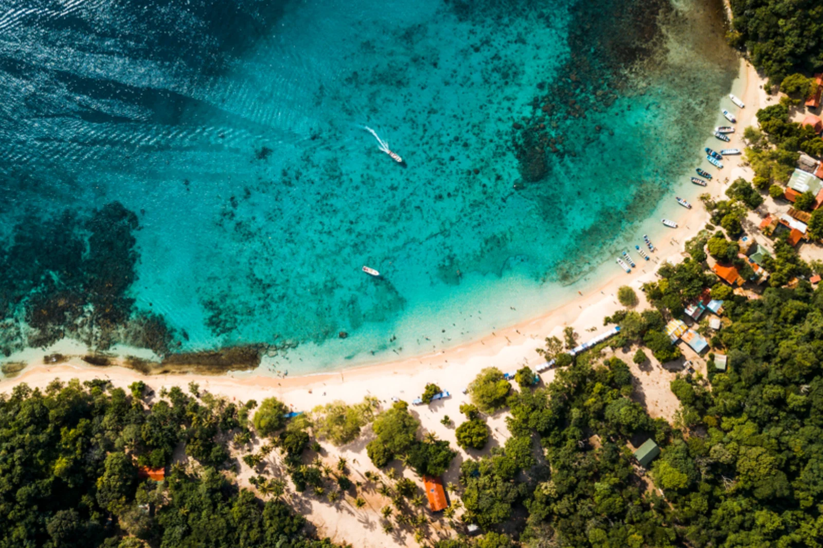 Vue zénithale de Playa Cristal avec une eau turquoise sur le récif, de petits bateaux et des kiosques au toit orange sous les arbres.