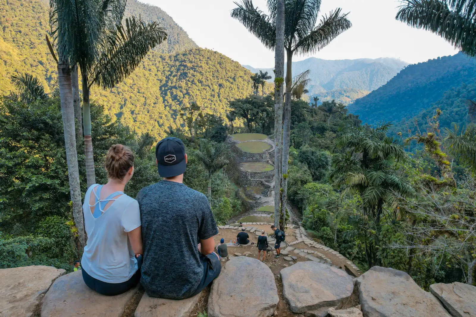 A couple sitting with their backs to the camera on a stone wall facing the terraces of Ciudad Perdida (Teyuna), with palms and forested mountains.