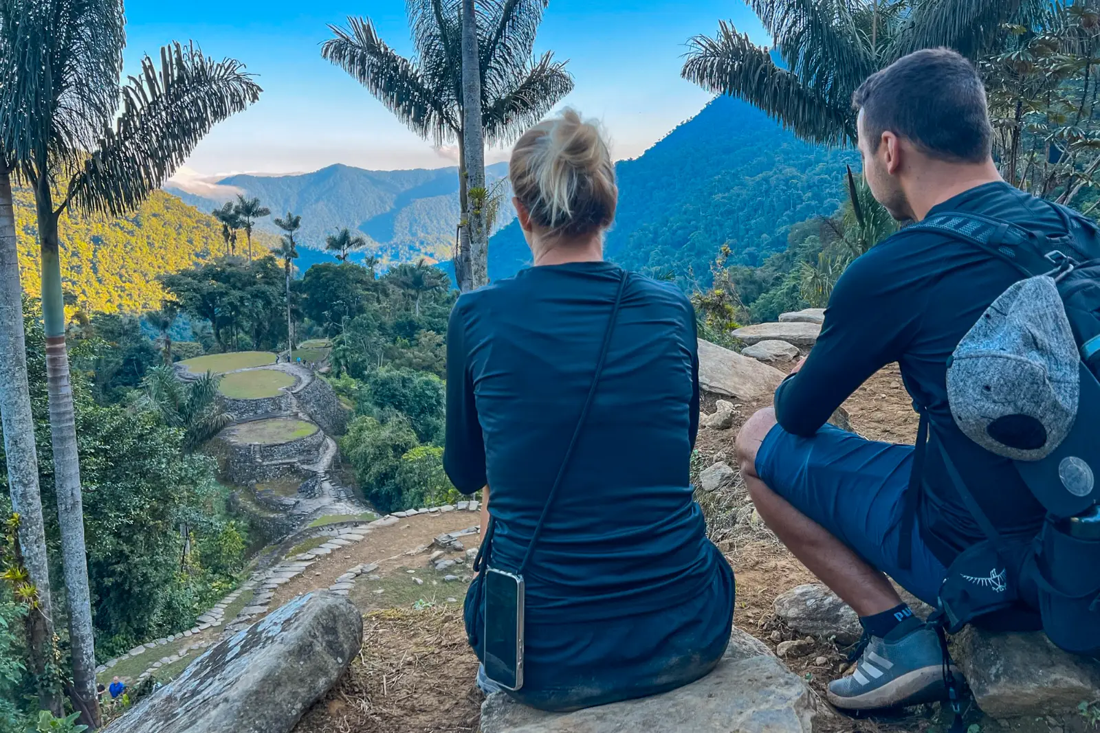 Two hikers seen from behind, one seated and one crouching with a backpack, look at the terraces of Ciudad Perdida (Teyuna) between tall palms.