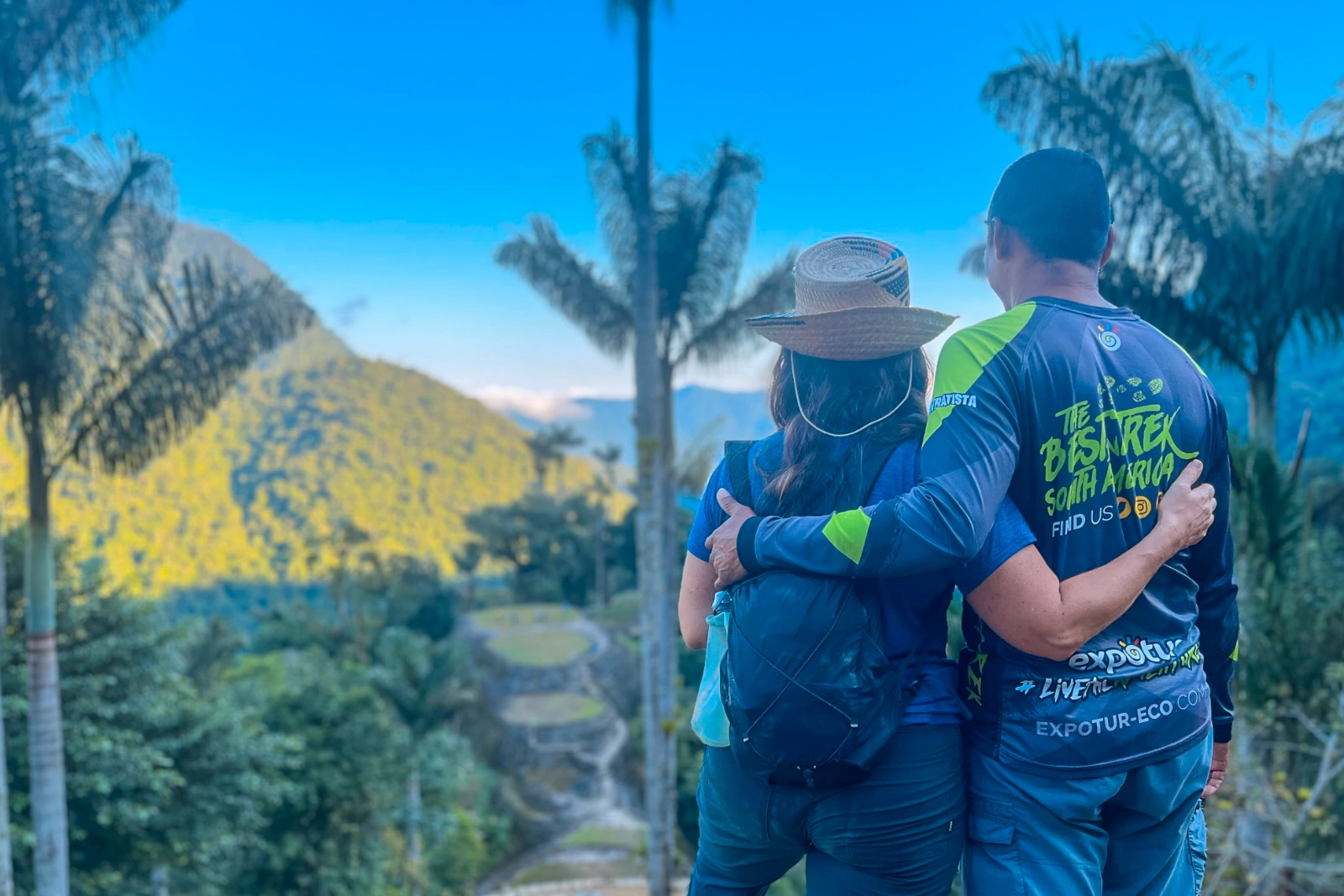 A couple seen from behind, arm in arm, facing the terraces of Ciudad Perdida (Teyuna) between palms, with a sunlit hillside beyond.