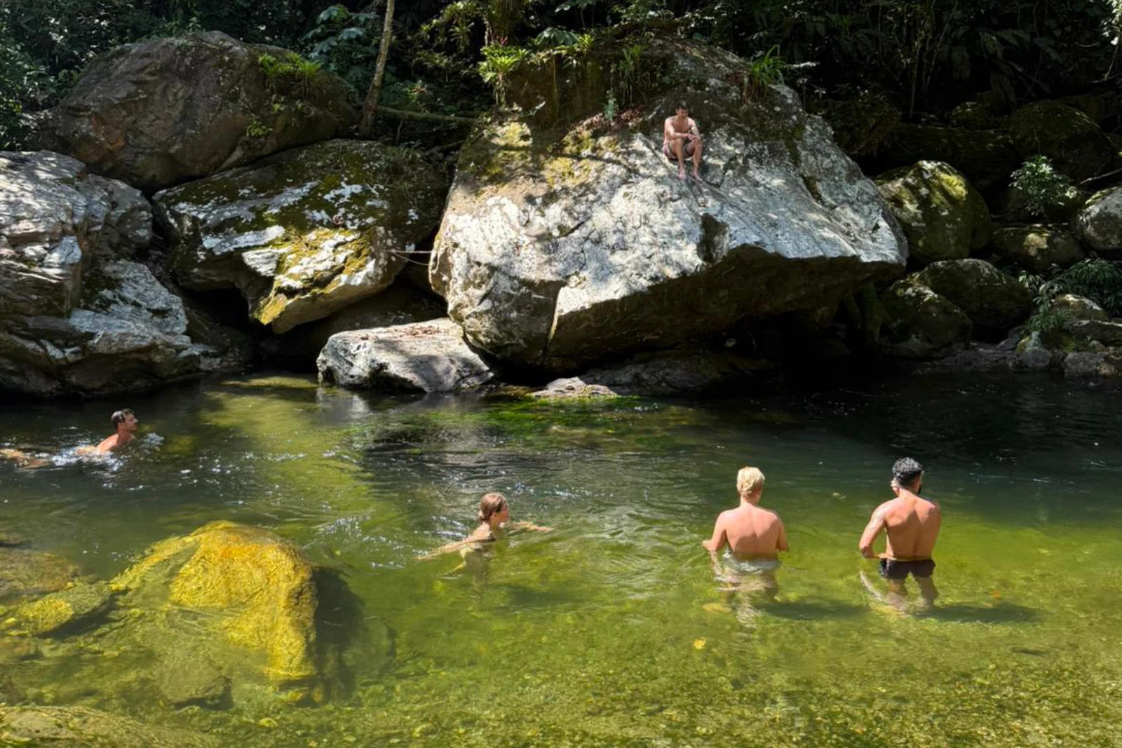 Des randonneurs se baignent dans un bassin d'eau verte et limpide d'une rivière, entre de gros rochers moussus, une personne assise sur un rocher.