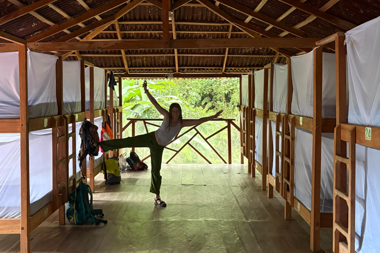 Randonneuse en posture de danse dans l'allée d'un dortoir en bois aux lits superposés et moustiquaires blanches, la forêt au fond.