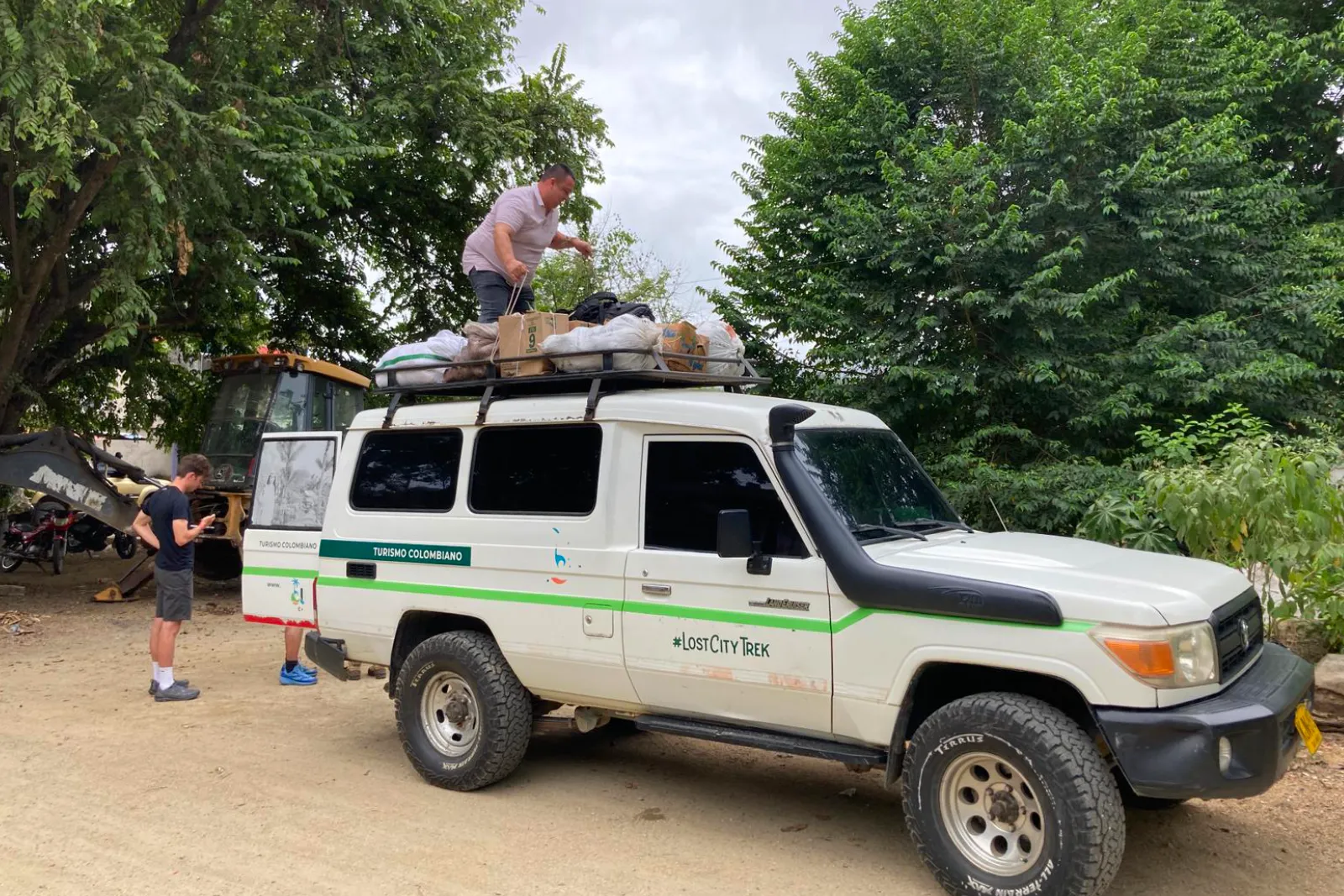 4×4 blanc au logo Lost City Trek sur la portière, bagages sur la galerie qu'un homme arrime depuis le toit, sous les arbres.