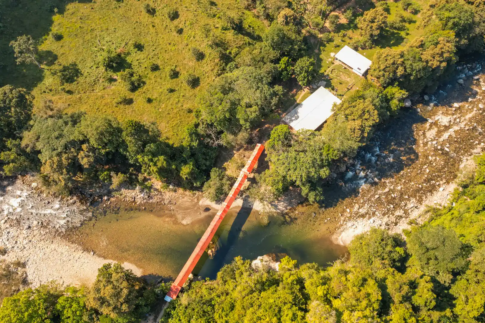 Aerial view of an orange suspension bridge over a shallow river in the forest, with two tin roofs on a grassy slope.