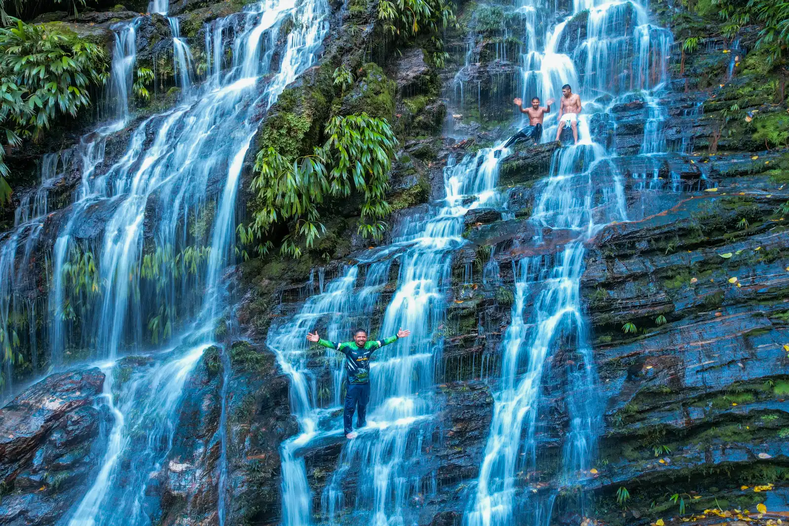 A wide waterfall cascading in several drops over dark rock with ferns, with two small figures standing in the water with arms open.