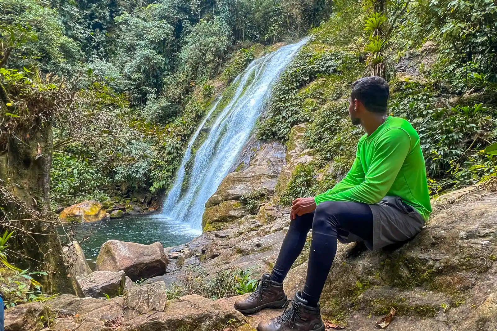 A hiker in a green shirt sits on a rock watching a waterfall drop into a pool in the forest, with large boulders on the bank.