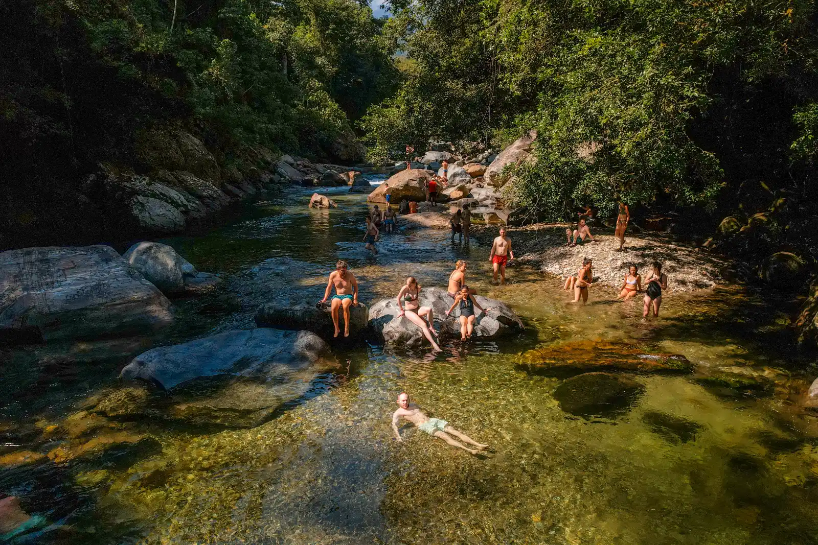 People swimming and sitting on the rocks of a clear pool in a river among large boulders, surrounded by forest.