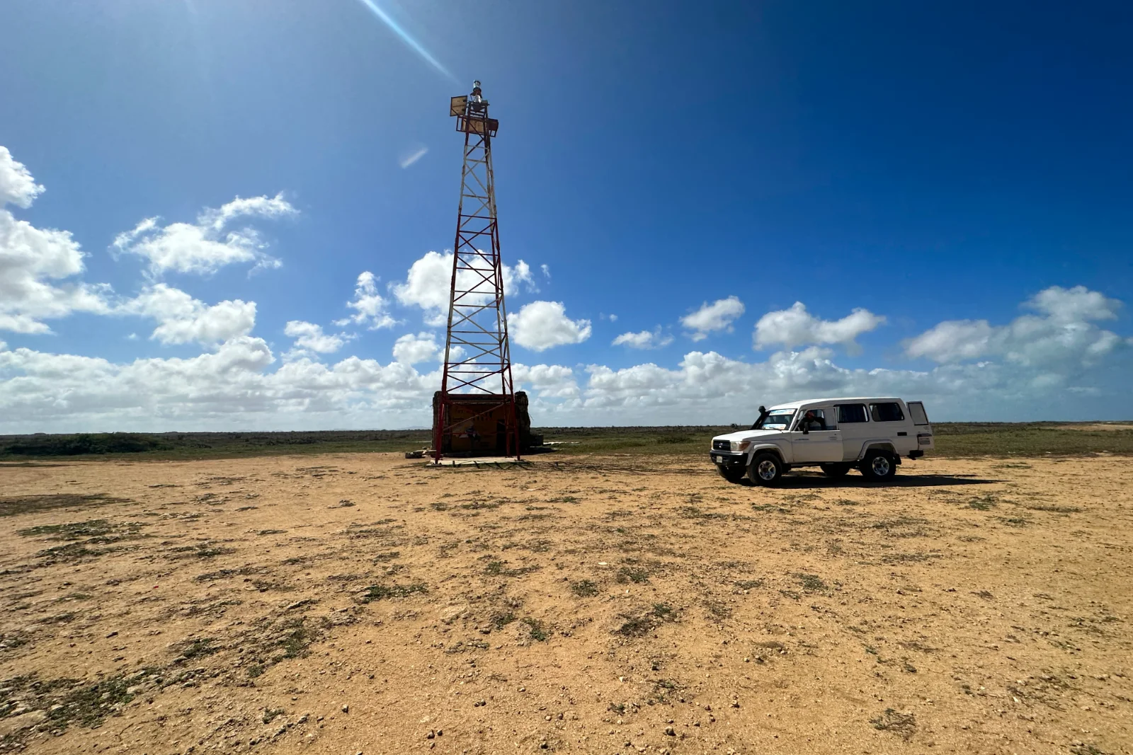 Tour en treillis rouge et blanc du phare de Punta Gallinas et un 4×4 blanc sur une plaine de sable, sous un ciel bleu nuageux.