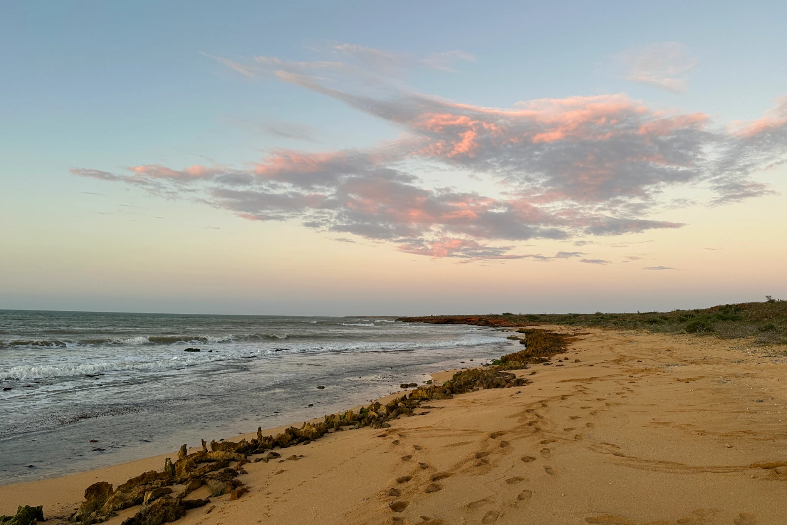 Plage de sable au crépuscule sous des nuages roses, rivage courbe bordé d'une ligne de rochers et traces de pas, à La Guajira.