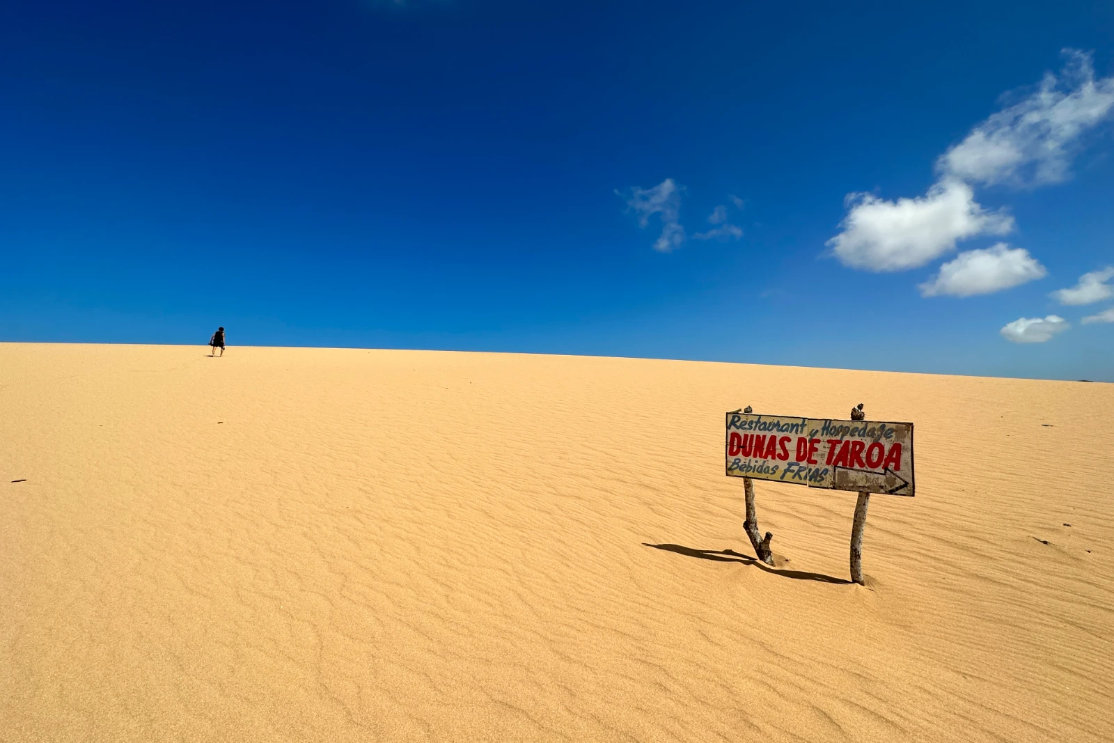 Panneau en bois des dunes de Taroa planté dans le sable, avec une personne au loin sur la crête et un ciel d'un bleu profond.