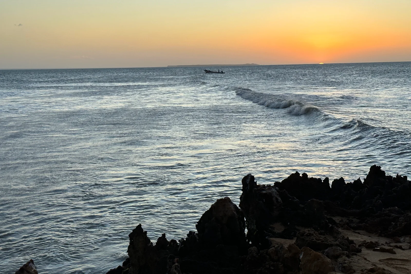 Coucher de soleil sur la mer avec une vague qui déferle, des rochers sombres et acérés au premier plan et une petite barque à l'horizon.