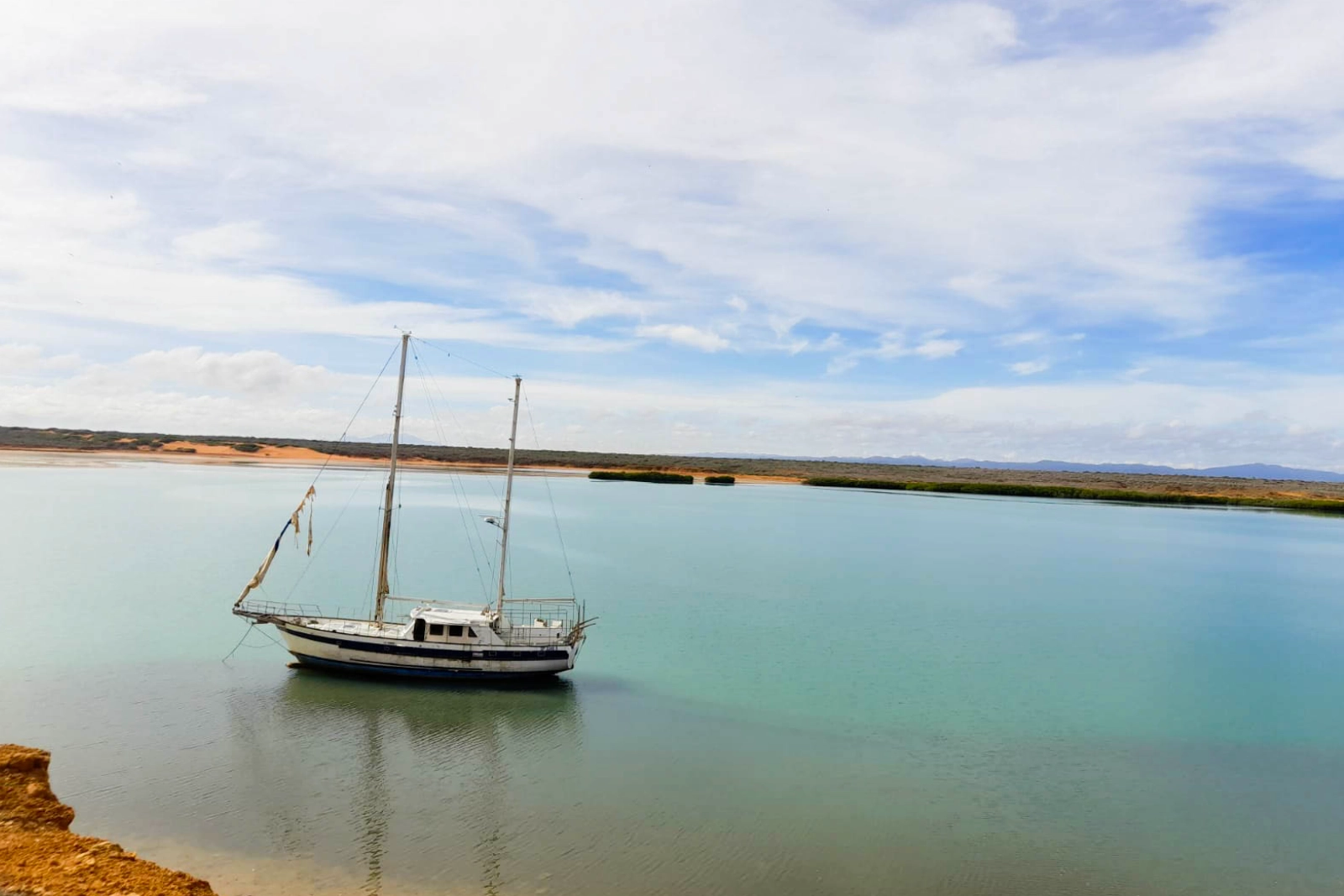 Voilier à deux mâts au mouillage dans l'eau verte et calme d'une baie de La Guajira, avec une rive basse de mangrove et un ciel nuageux.