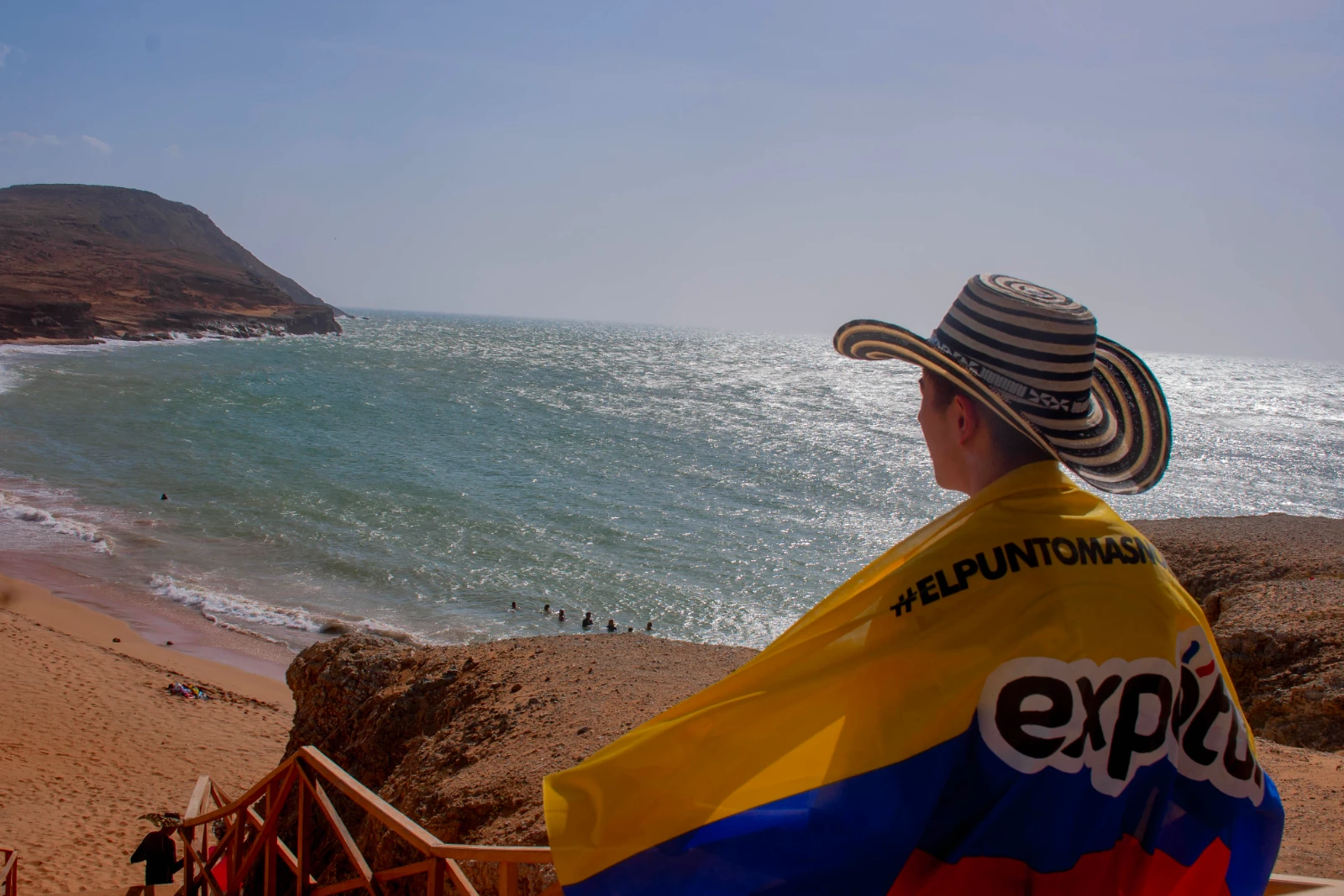 Personne de dos coiffée d'un sombrero vueltiao, un drapeau colombien au logo d'Expotur sur les épaules, face à la mer depuis une falaise.