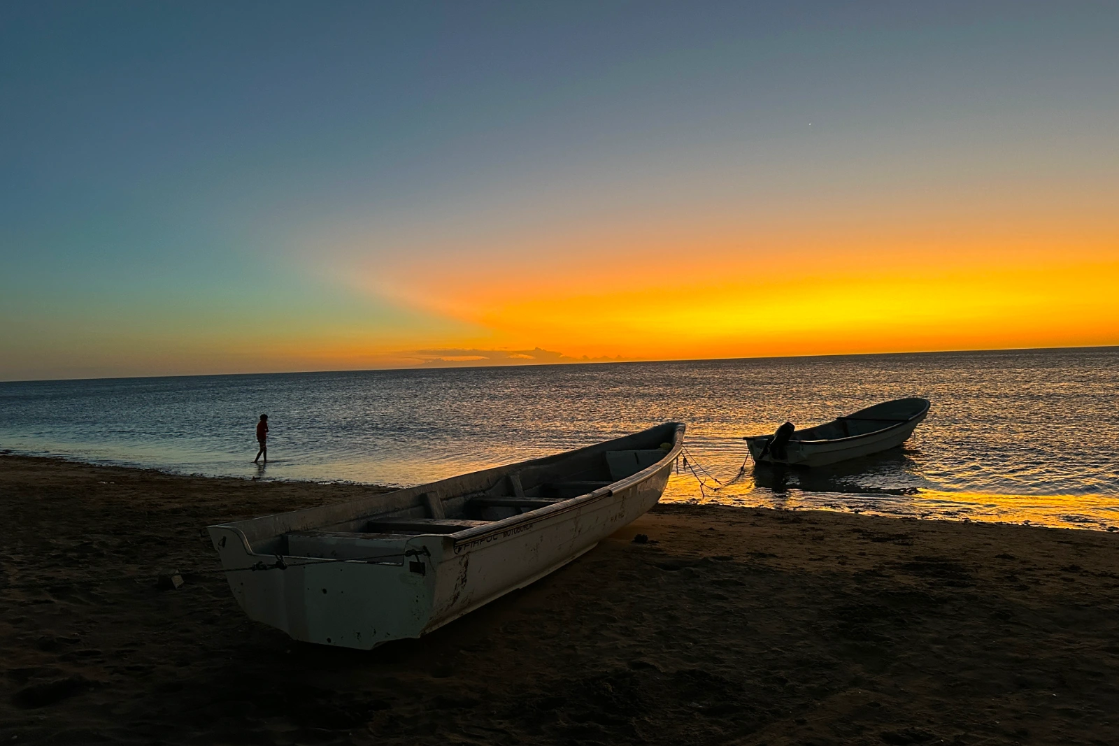 Deux barques en bois sur la plage au coucher du soleil, ciel orange sur la mer et silhouette lointaine d'une personne sur le rivage.
