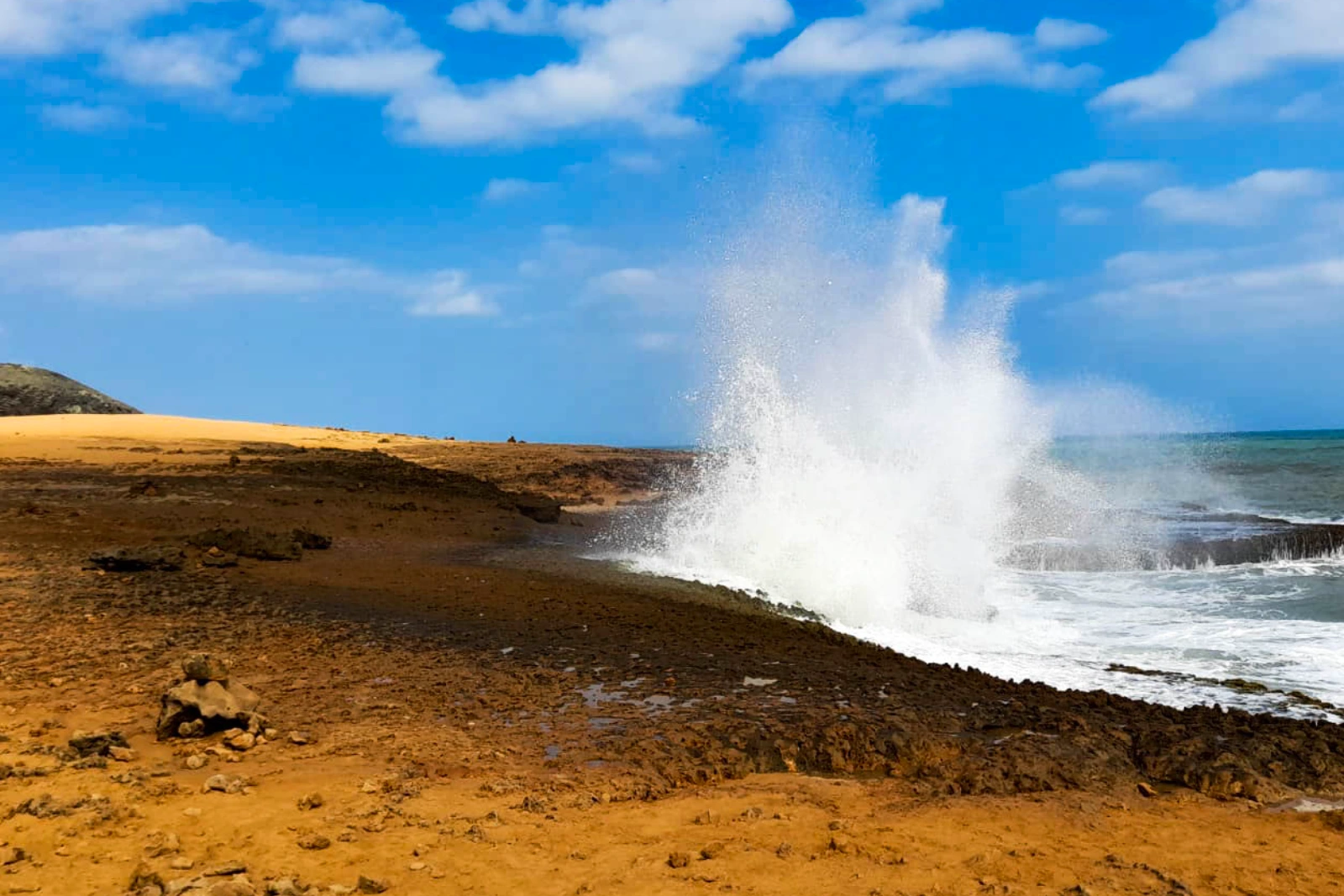 Vague qui se brise et jaillit haut contre une côte de roche ocre, près d'une plage de sable, sous un ciel bleu nuageux, à La Guajira.