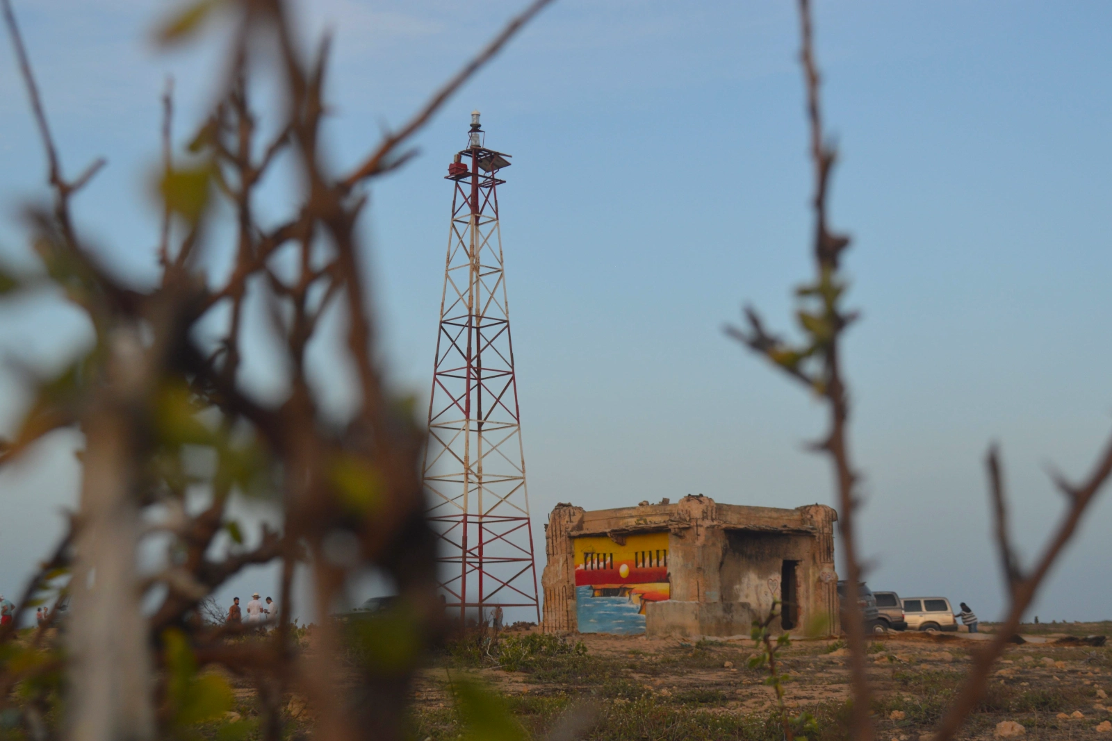 Tour métallique rouge et blanc du phare de Punta Gallinas et la cabane peinte à côté, vues entre des branches sèches au crépuscule.