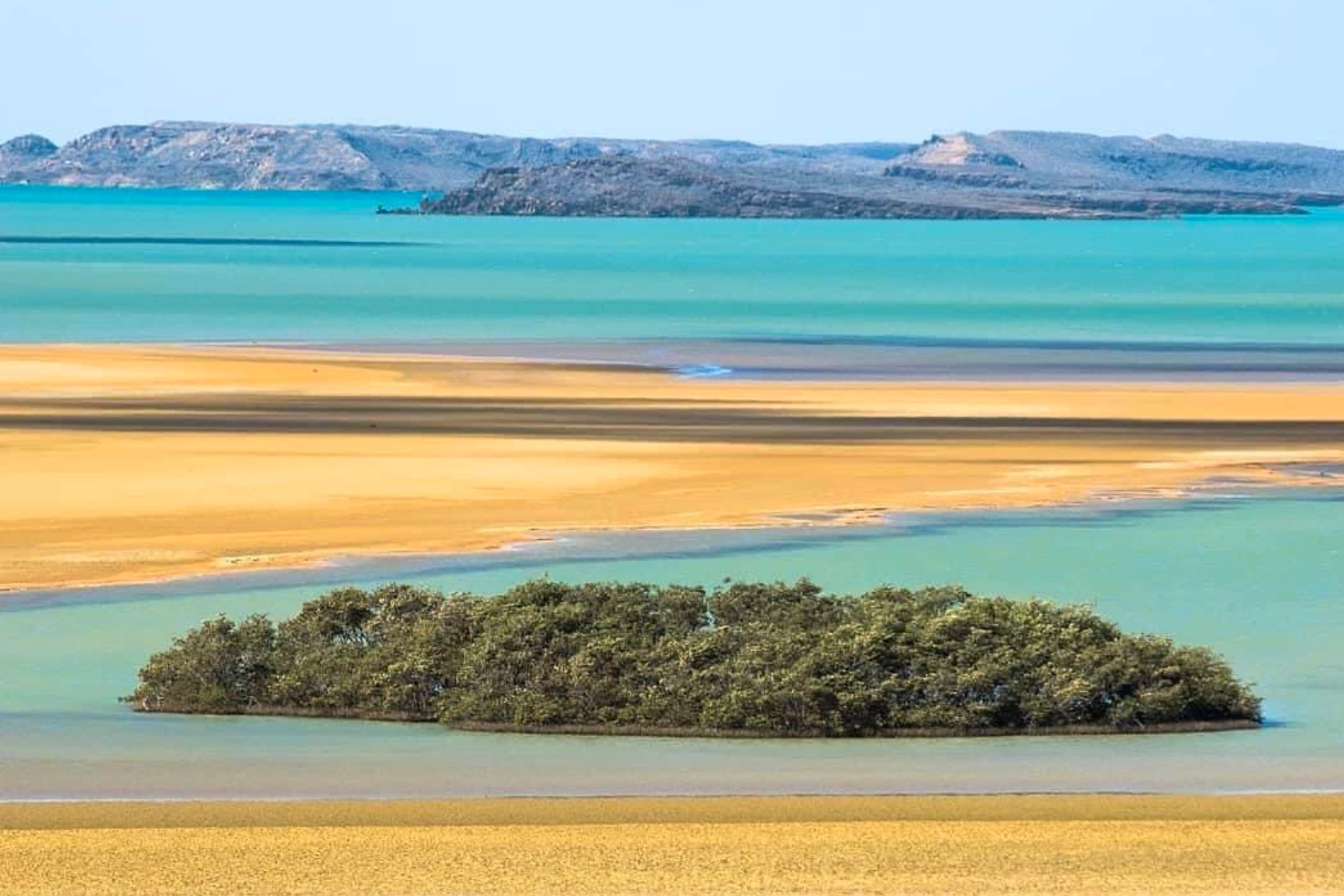 Bancs de sable ocre entre des eaux turquoise peu profondes, un îlot de mangrove au premier plan et des collines basses dans la brume.
