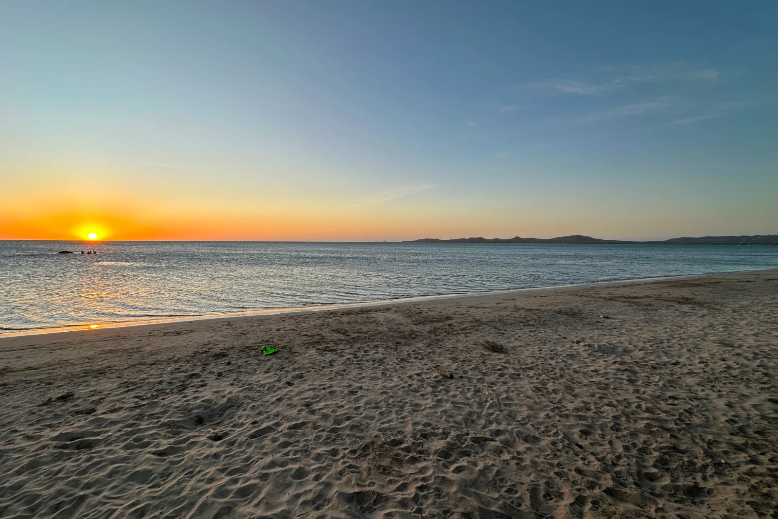 Coucher de soleil sur la mer vu d'une plage de sable marquée de traces de pas, soleil sur l'horizon, eau calme et terre basse au loin.