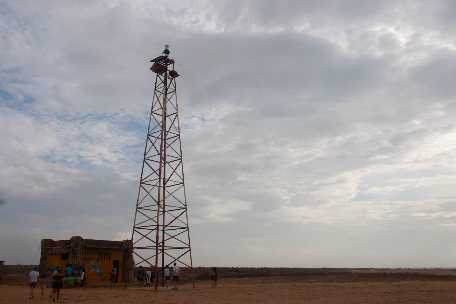 Tour en treillis du phare de Punta Gallinas et une cabane en pierre sur une plaine, sous un ciel couvert, avec de petits visiteurs au pied.