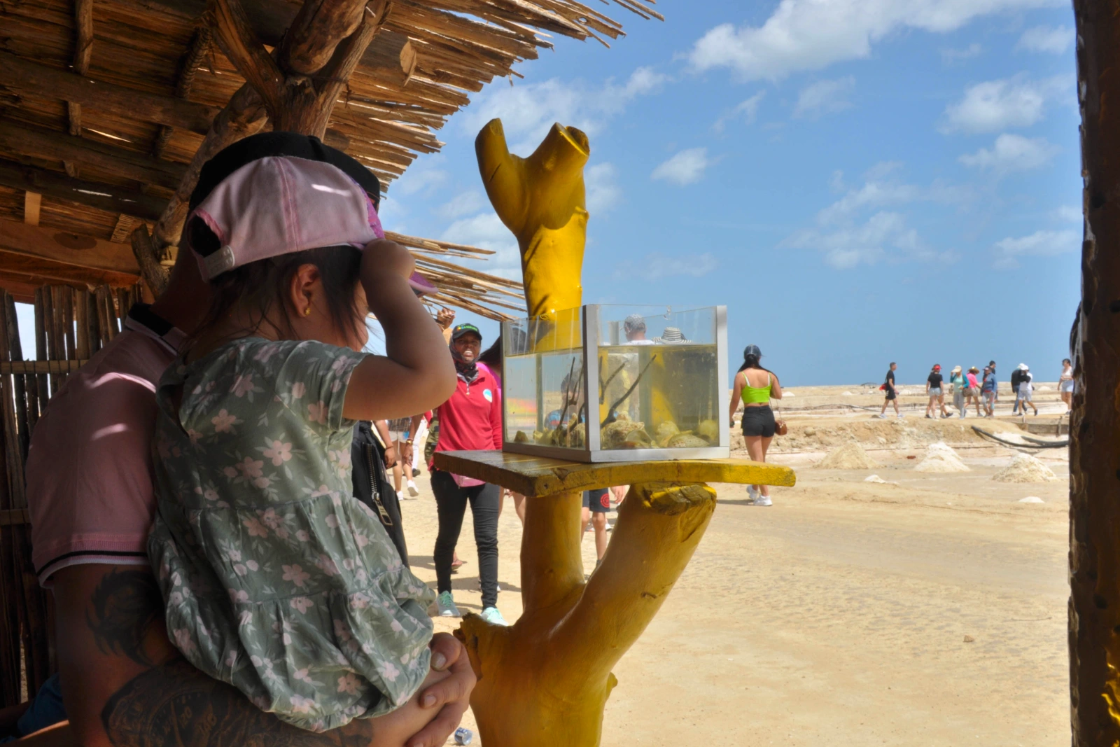Un voyageur portant une fillette sous un toit de palmes, près d'un support en bois jaune avec une boîte vitrée, et une plage claire au fond.