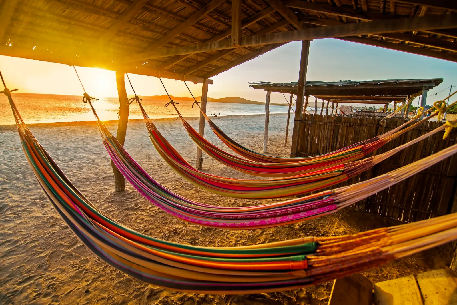 Chinchorros (hamacs) colorés suspendus sous une enramada face à la mer au coucher du soleil, soleil bas à gauche, à Cabo de la Vela.