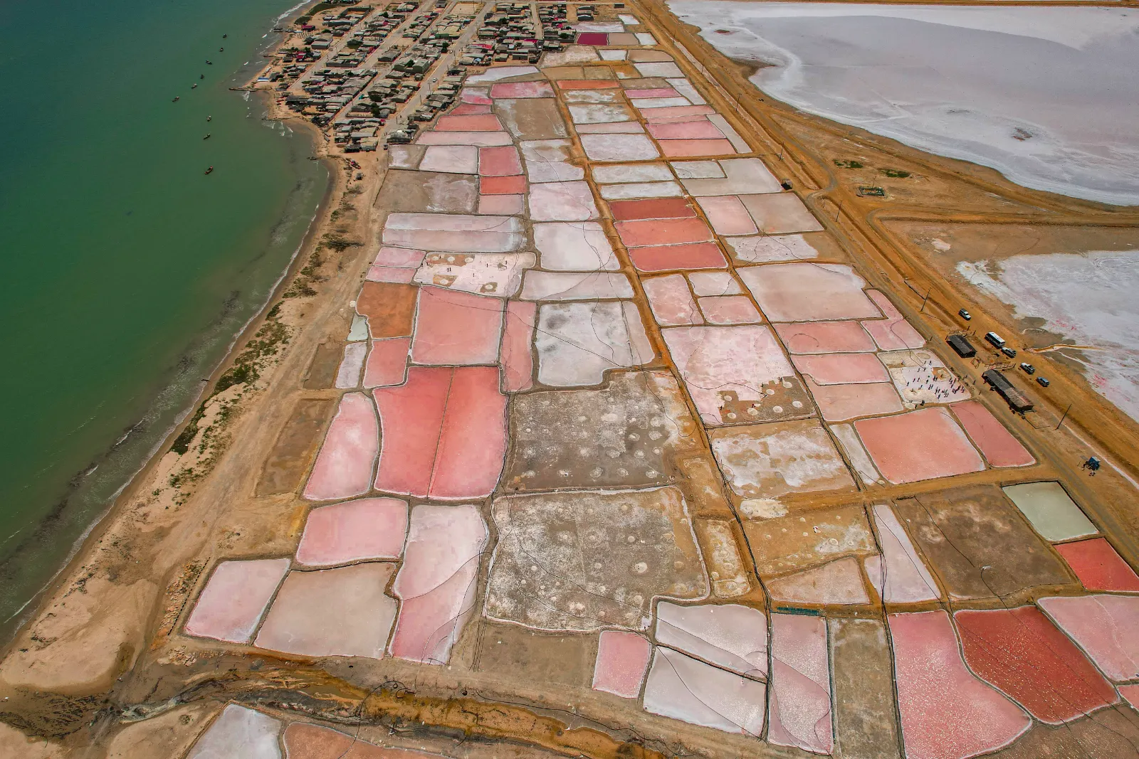 Vue aérienne des bassins roses et blancs des salines de Manaure au bord de la mer, avec la ville en haut et des camions sur la route.